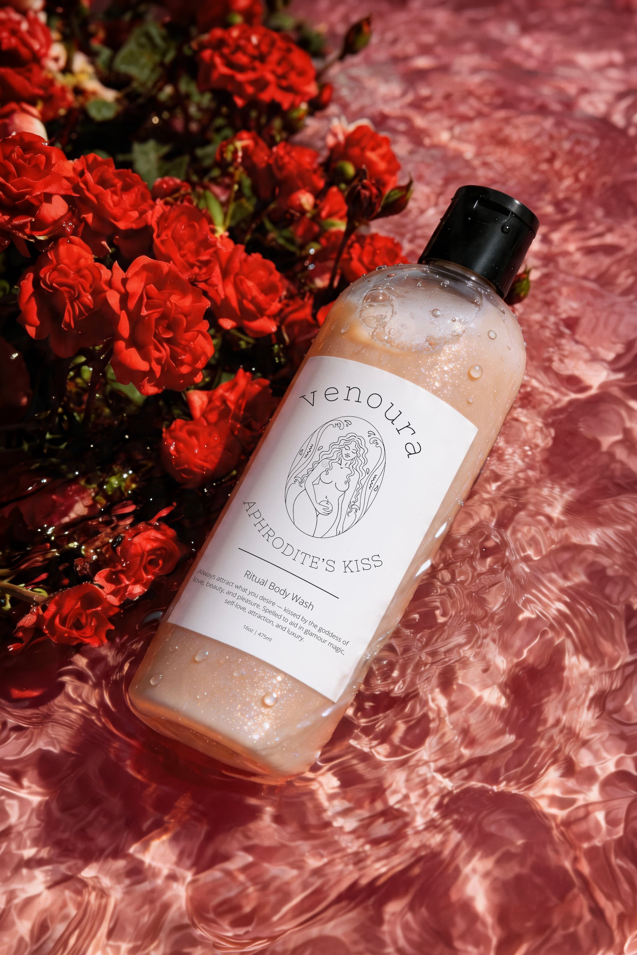 Venoura Kierodite's Kiss Soak Body Wash in a clear bottle with a black cap, resting on pink water with red roses in the background.