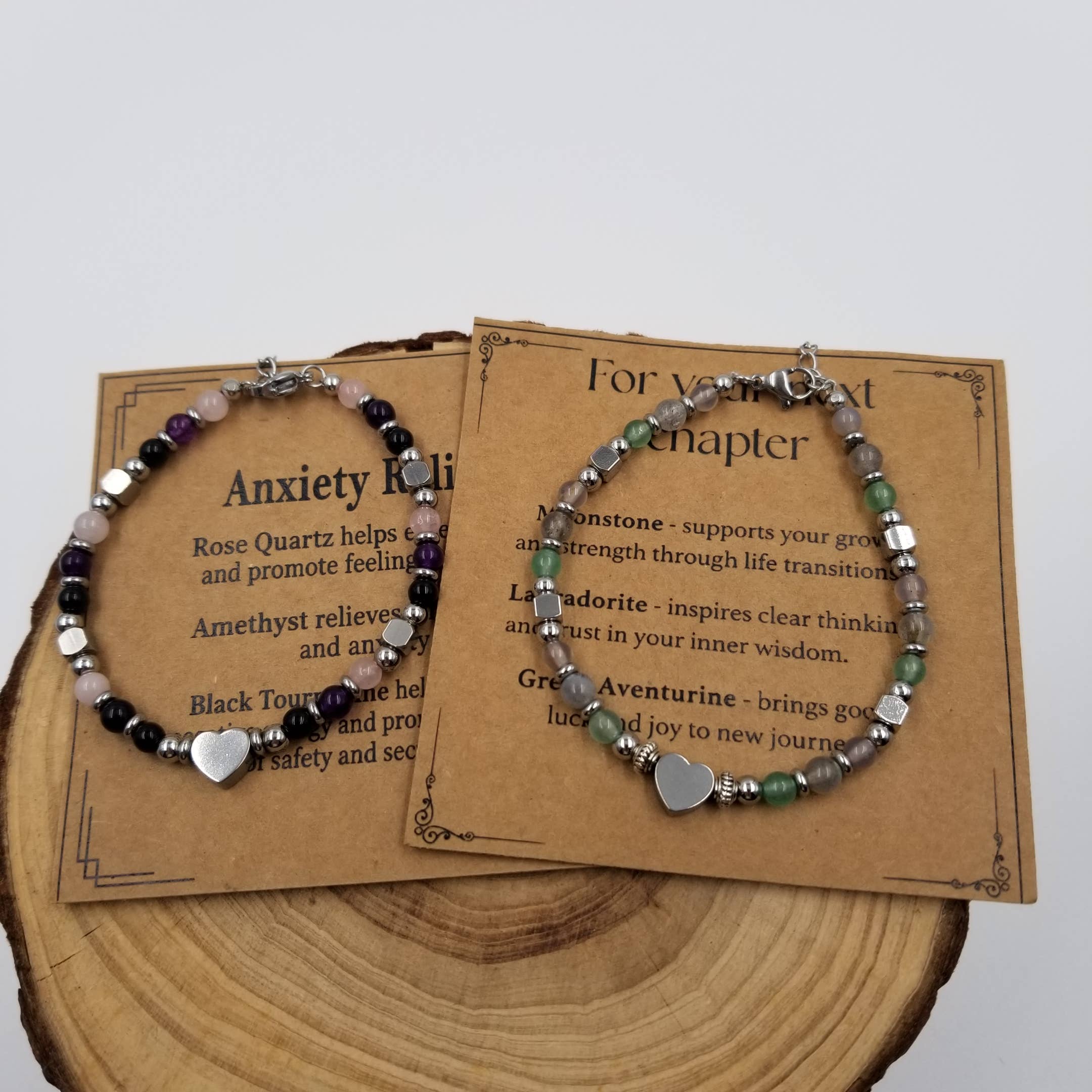 Two beaded anxiety relief bracelets displayed on brown printed cards over a wood slice. Each bracelet has mixed gemstone beads in black, purple, pink, green, and silver, with a silver heart charm.