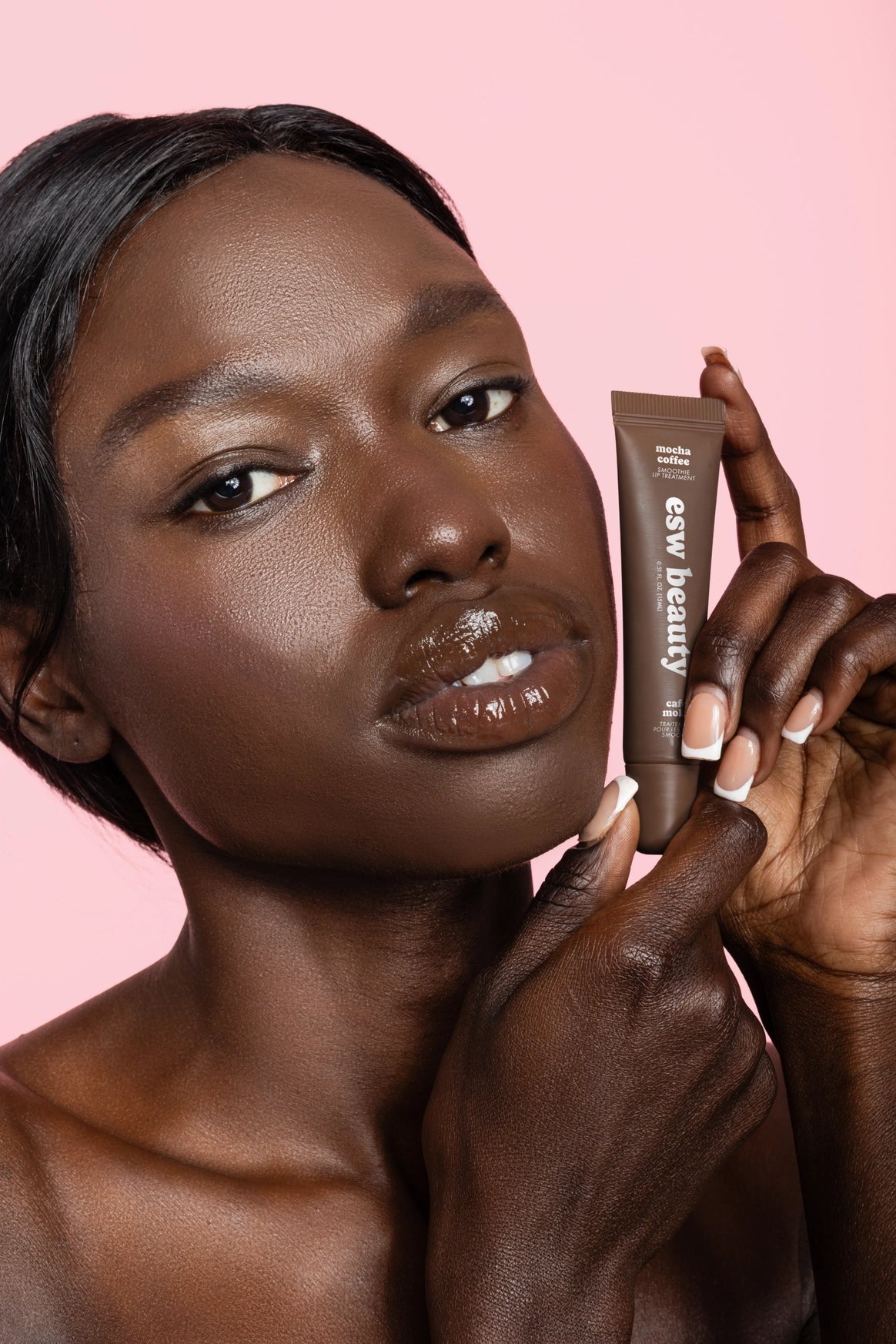 A close-up of a model with dark skin holding a brown cosmetic tube labeled 'css beauty' against a solid pink background.
