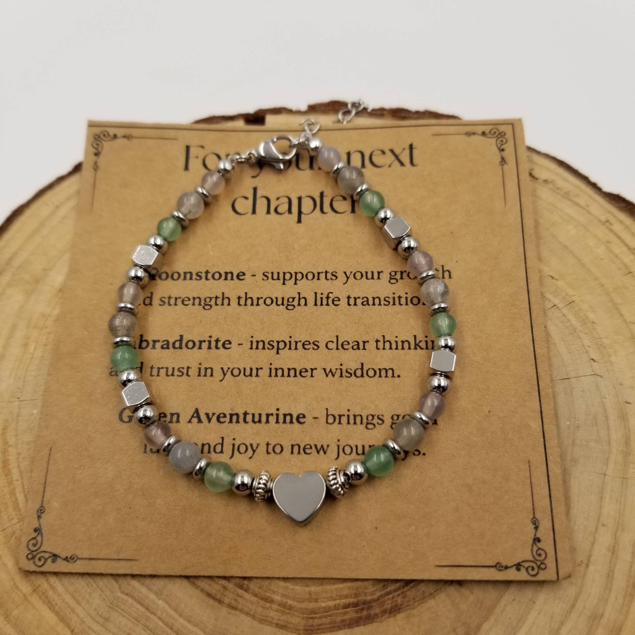 A beaded bracelet with gray agate-like beads, green beads, silver-toned spacer beads, and a heart-shaped silver charm, displayed on a brown kraft card over a wood slice background.