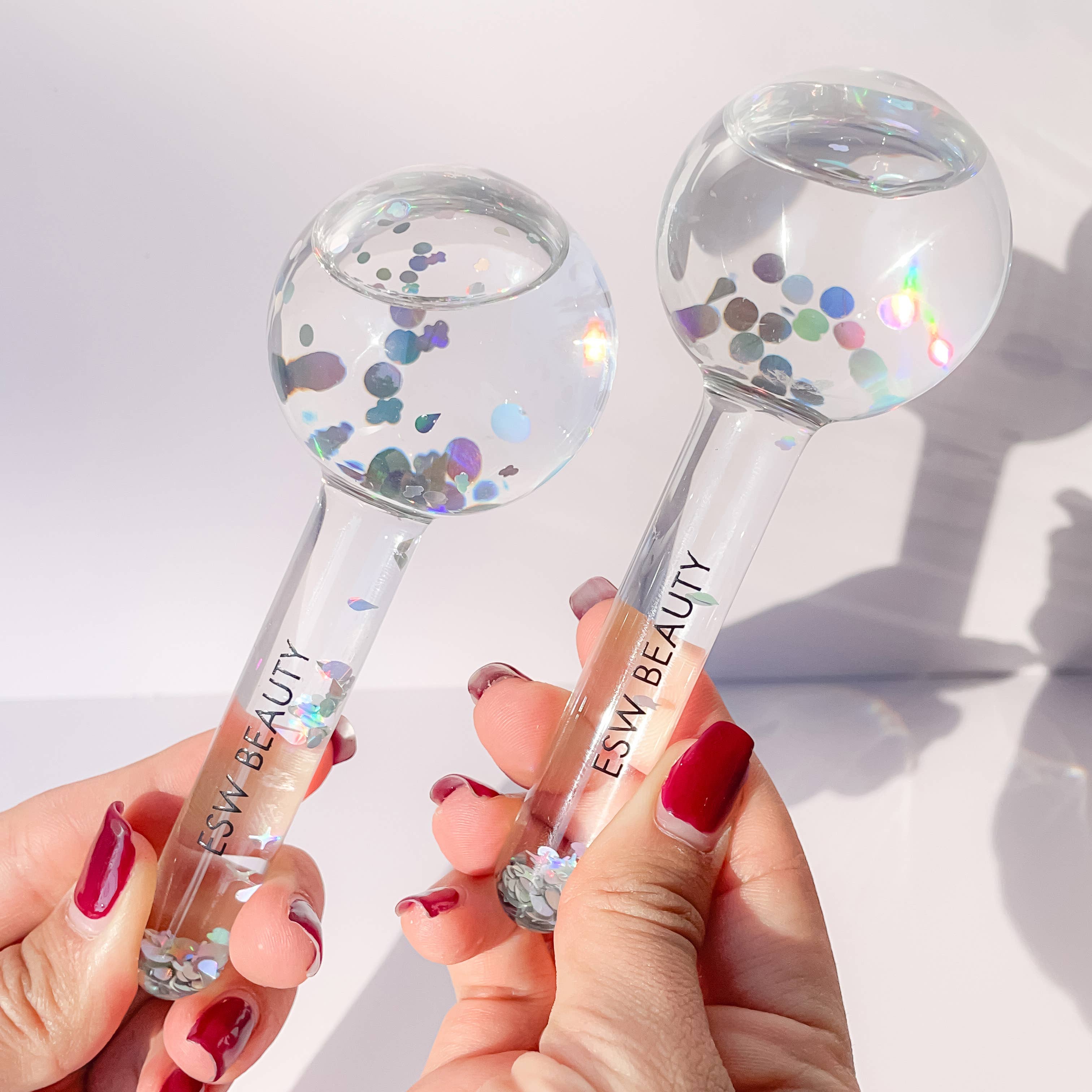 Two clear beauty tools with colorful confetti inside, held by a person with red nail polish on a light background.