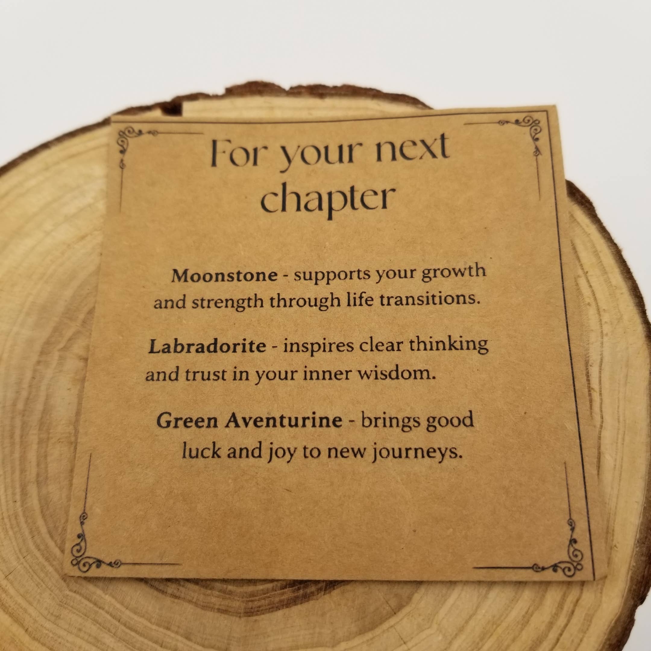 A square kraft-paper card with black decorative corner flourishes and the text 'For your next chapter' followed by three short phrases, placed on a round natural wood slice against a light neutral background.