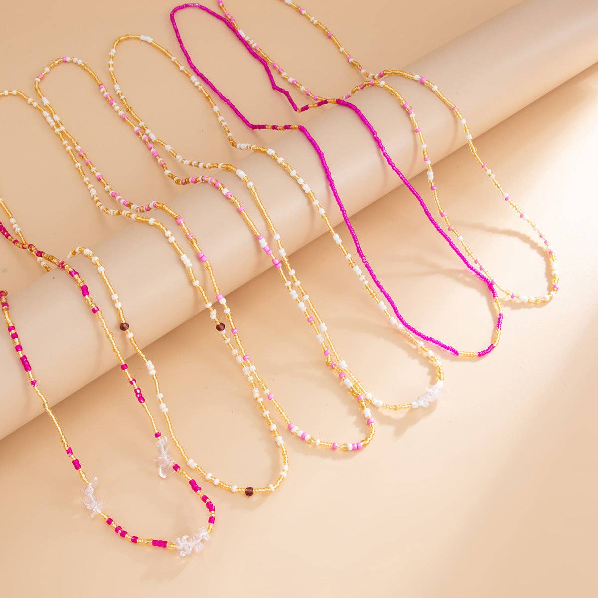 Five delicate beaded necklaces with gold-tone chains and mixed bead colors, including white pearls, pink, purple, and red beads, arranged on a beige background.