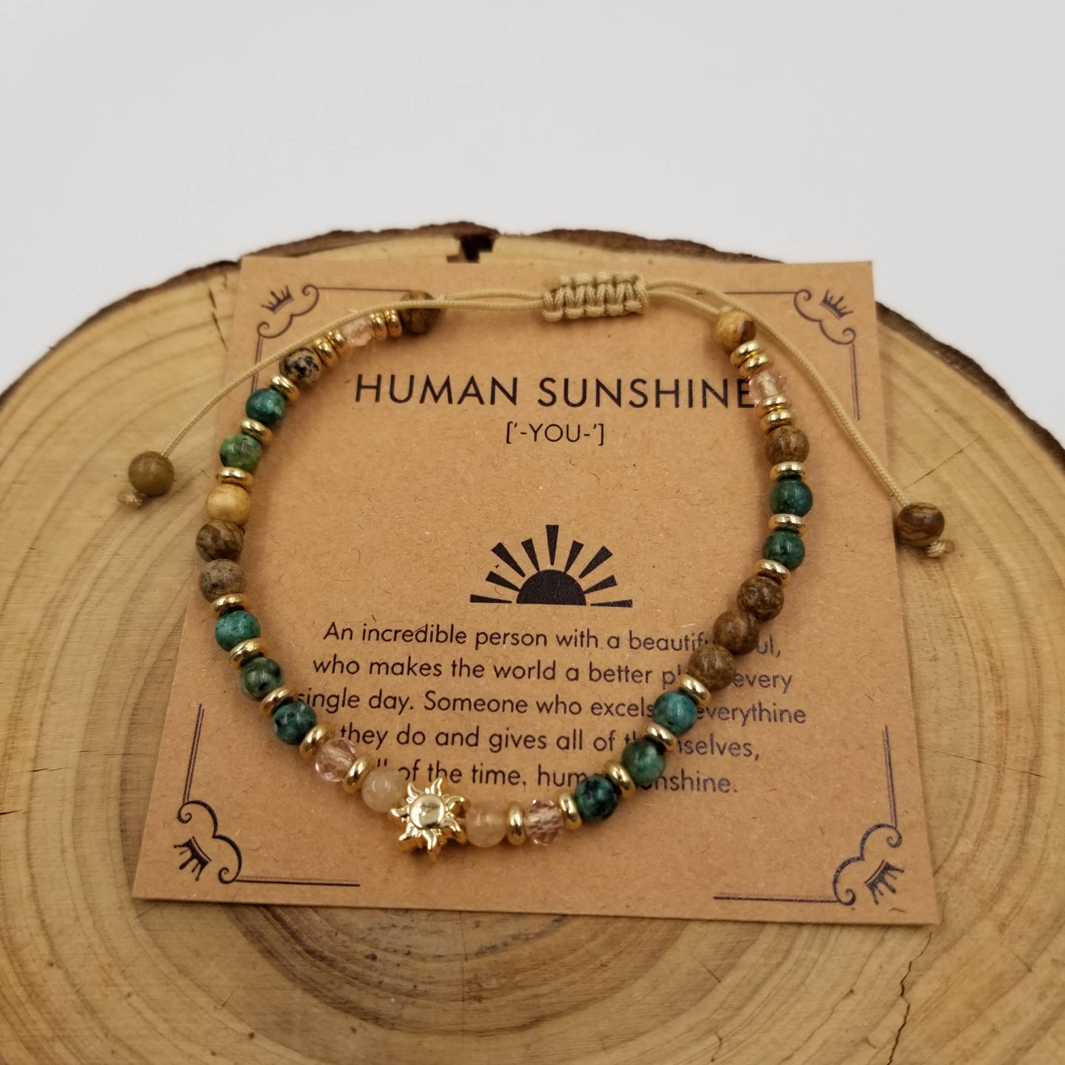 A beaded cord bracelet displayed on a kraft card that reads 'HUMAN SUNSHINES! [YOU-]', placed on a round wood slice. The bracelet has mixed earthy beads in green, brown, beige, and gold-toned accents, including a small gold sun charm at the front.
