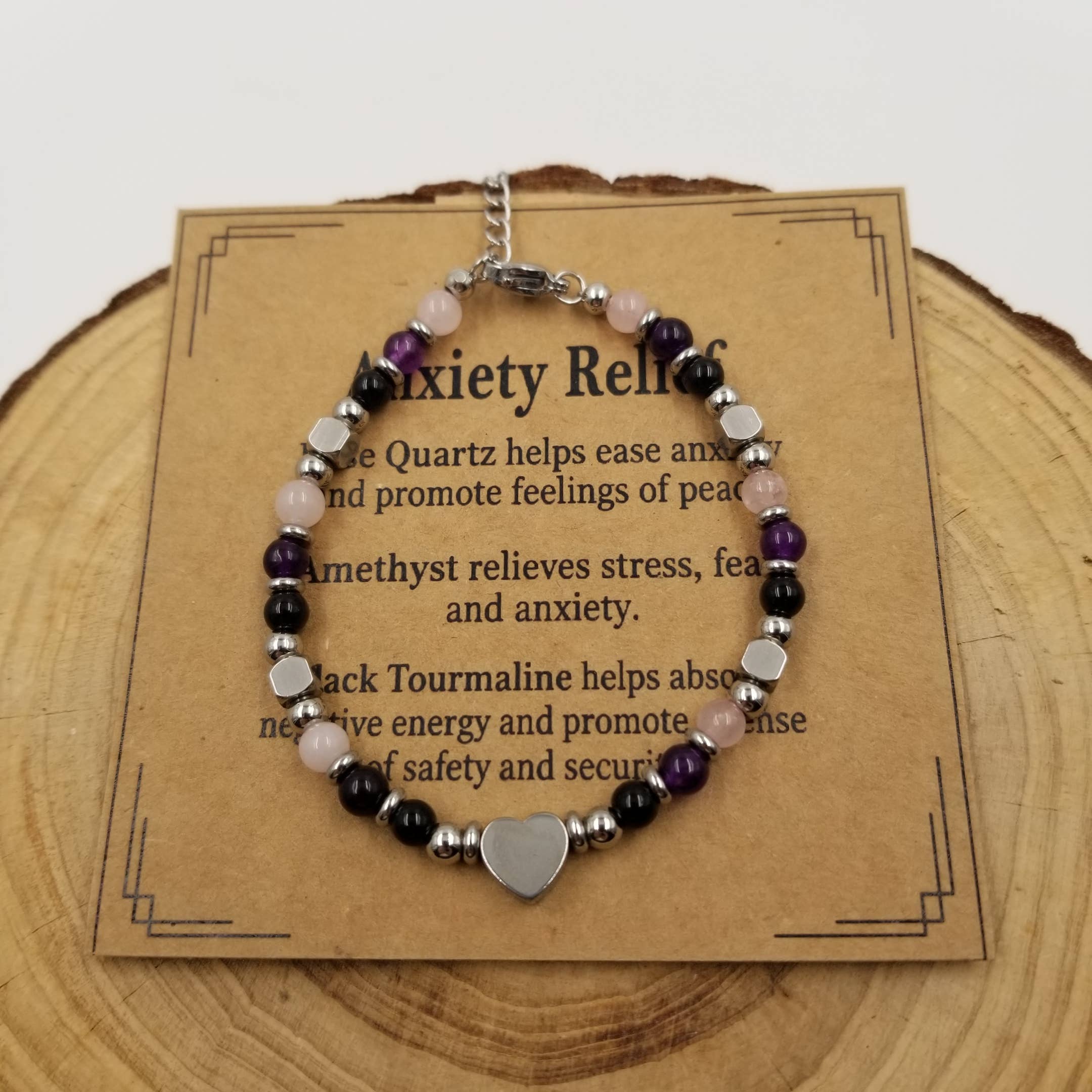 An anxiety relief beaded bracelet displayed on a kraft paper card, which is placed on a rustic wood slice. The bracelet features purple amethyst, pink rose quartz, black tourmaline, silver spacer beads, and a central silver heart charm.