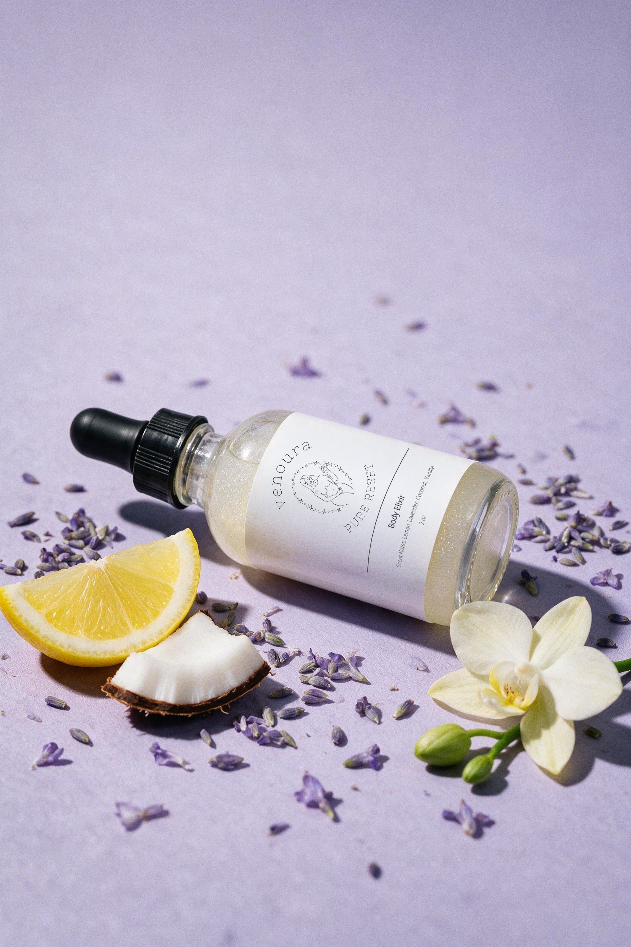 A glass dropper bottle of body wash with a white label lies on a lavender surface, surrounded by scattered dried lavender buds, a lemon wedge, a coconut half, and a white orchid flower.