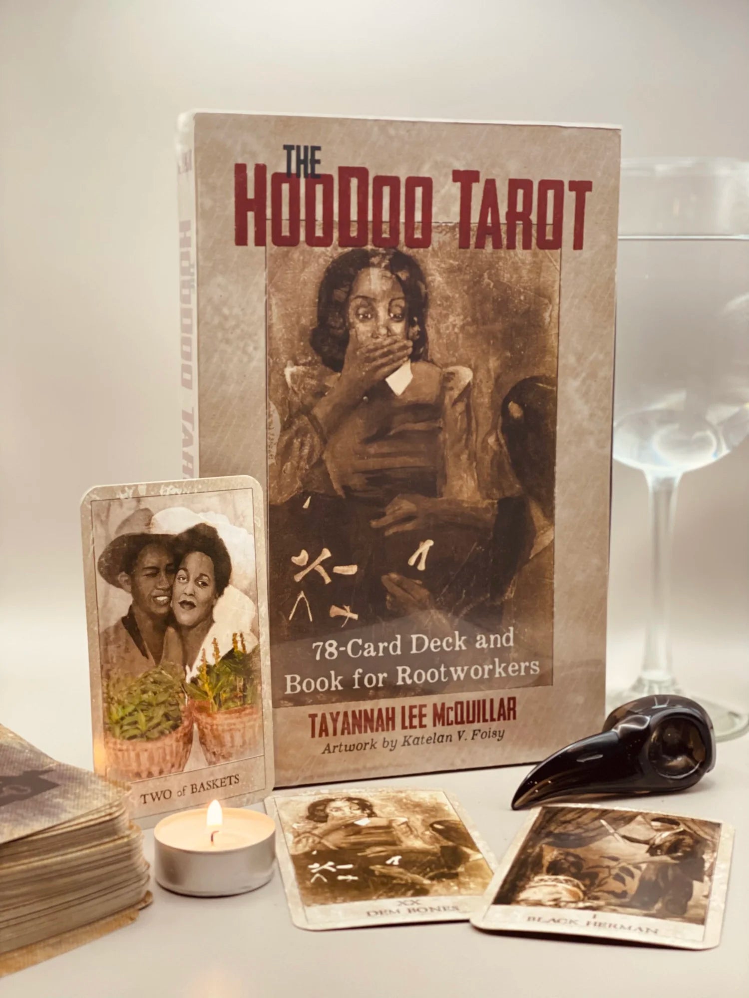 The Hoodoo Tarot deck and book set displayed on a tabletop with tarot cards, a lit candle, a glass, and a black bird skull against a neutral light background.