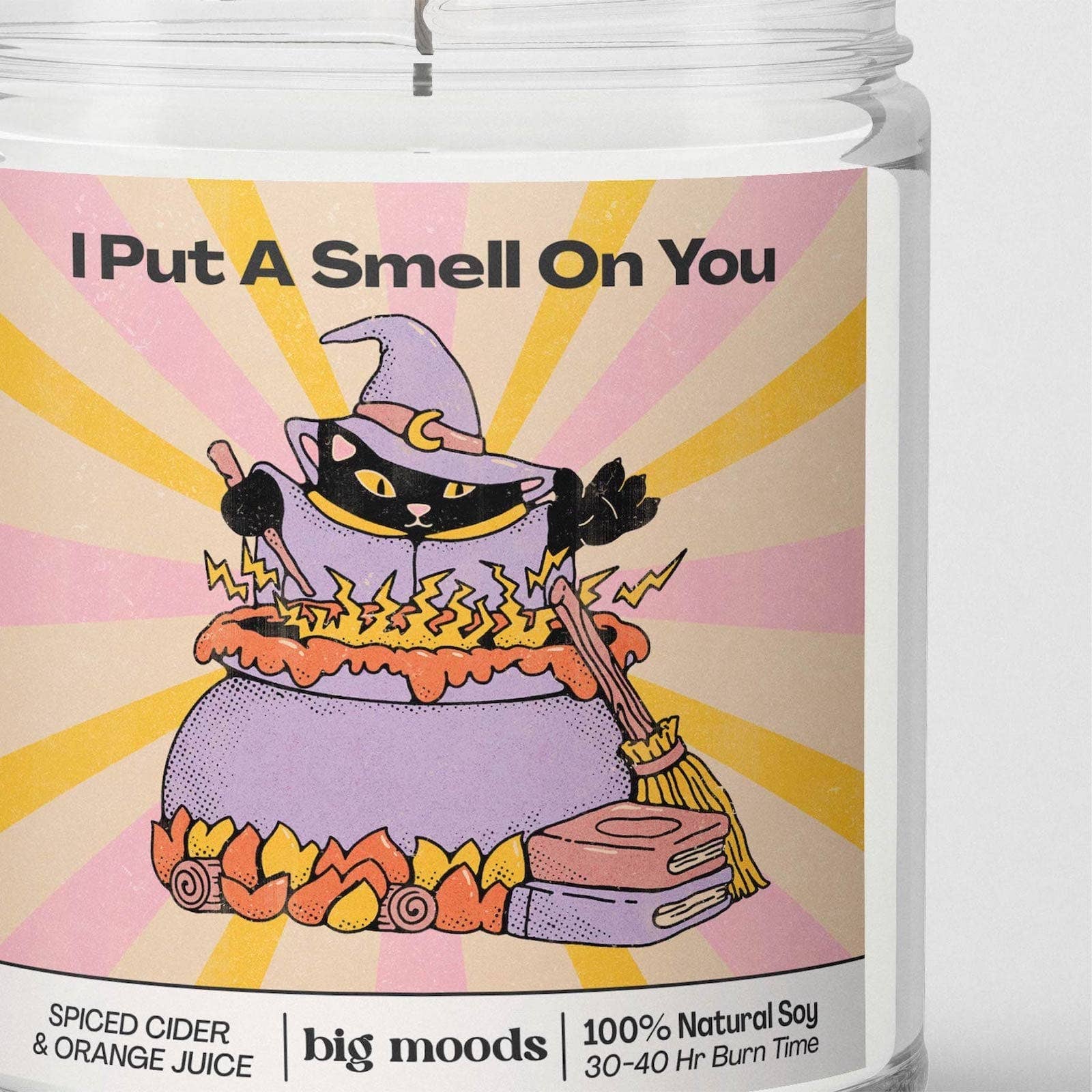 A close-up of a candle jar label with the scent name 'I Put A Smell On You' and an illustration of a black cat in a witch hat stirring orange liquid in a cauldron. The label also shows 'big moods', '100% Natural Soy', and 'Spiced Cider & Orange Juice'.