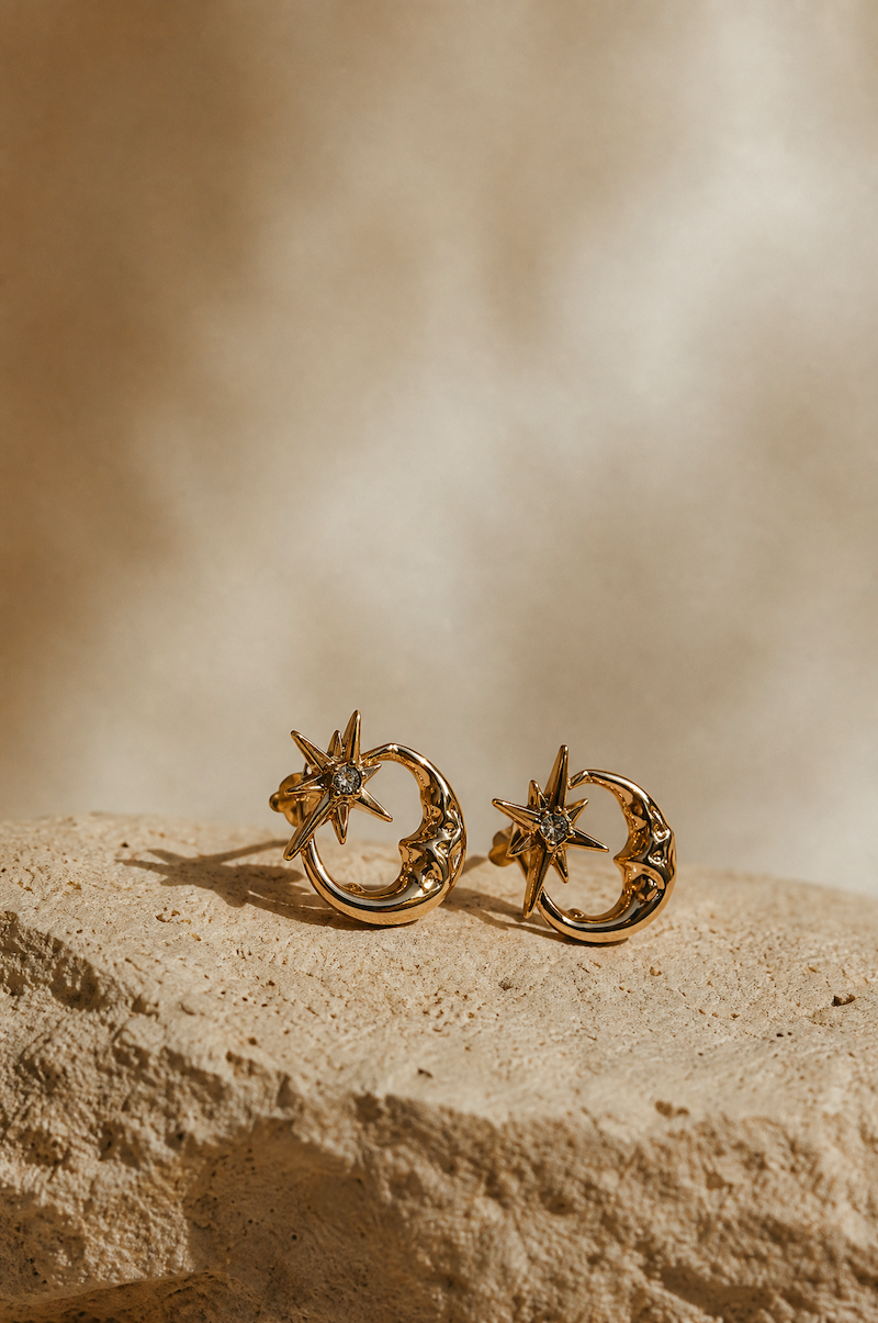 A pair of gold crescent moon and star stud earrings resting on a textured beige stone surface with a warm, sunlit beige background.