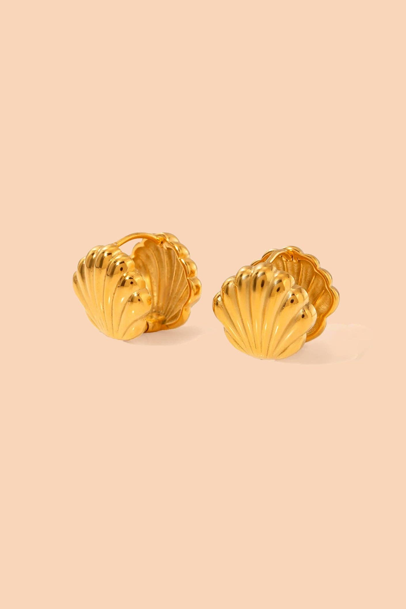 A pair of gold shell-shaped stud earrings with a textured, ribbed surface, displayed on a solid pale peach background.