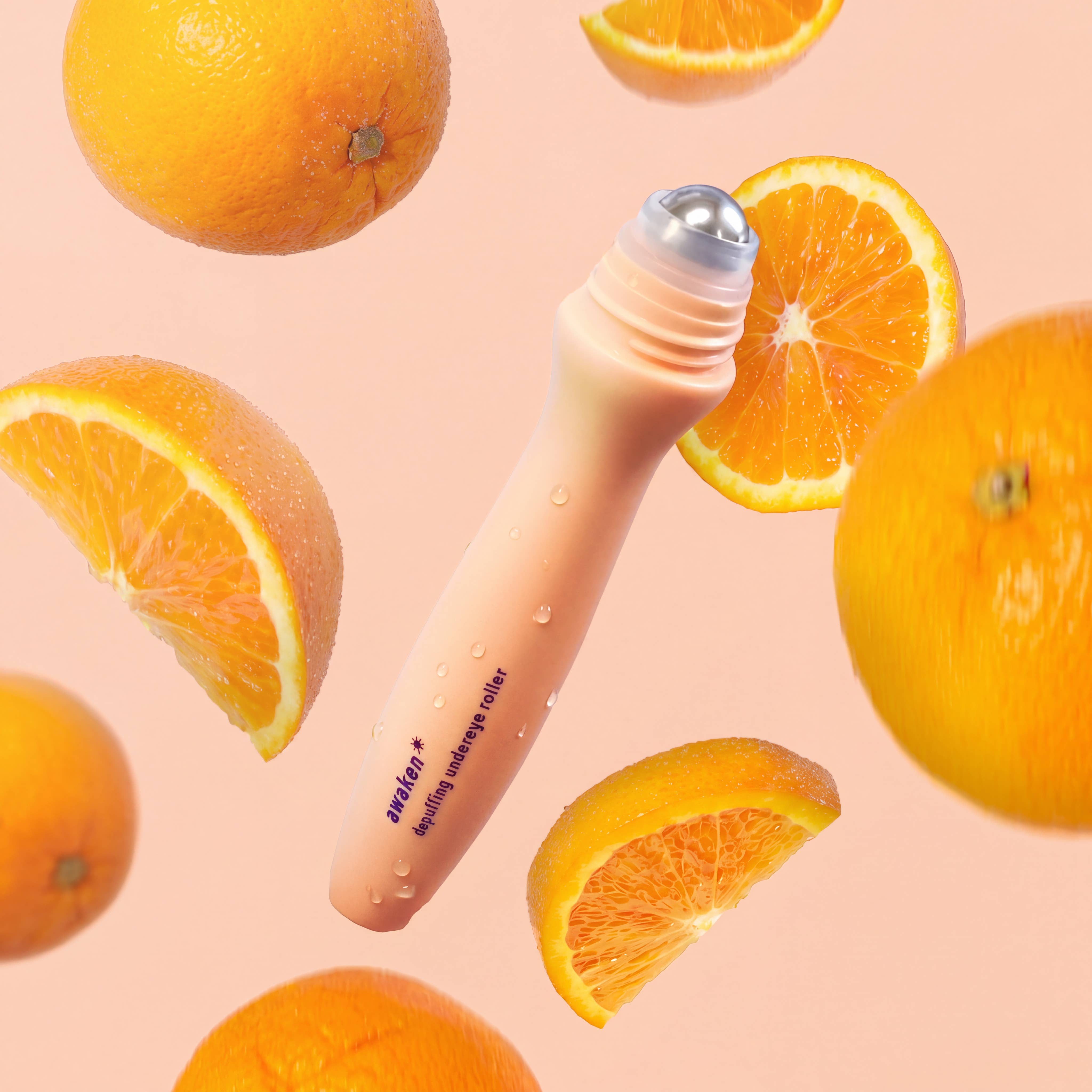 A peach-colored cosmetic rollerball bottle labeled 'amjies' and 'spiny oregano mask' is centered against a soft peach background, surrounded by floating whole oranges and orange slices.