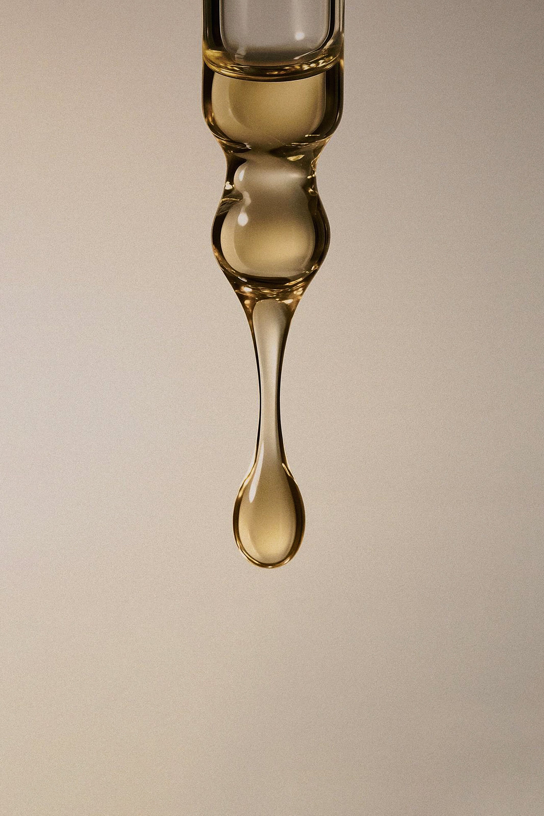 A close-up studio shot of a clear glass dropper tube with a bulbous middle section, from which a single glossy amber-yellow liquid drop hangs against a warm beige background.