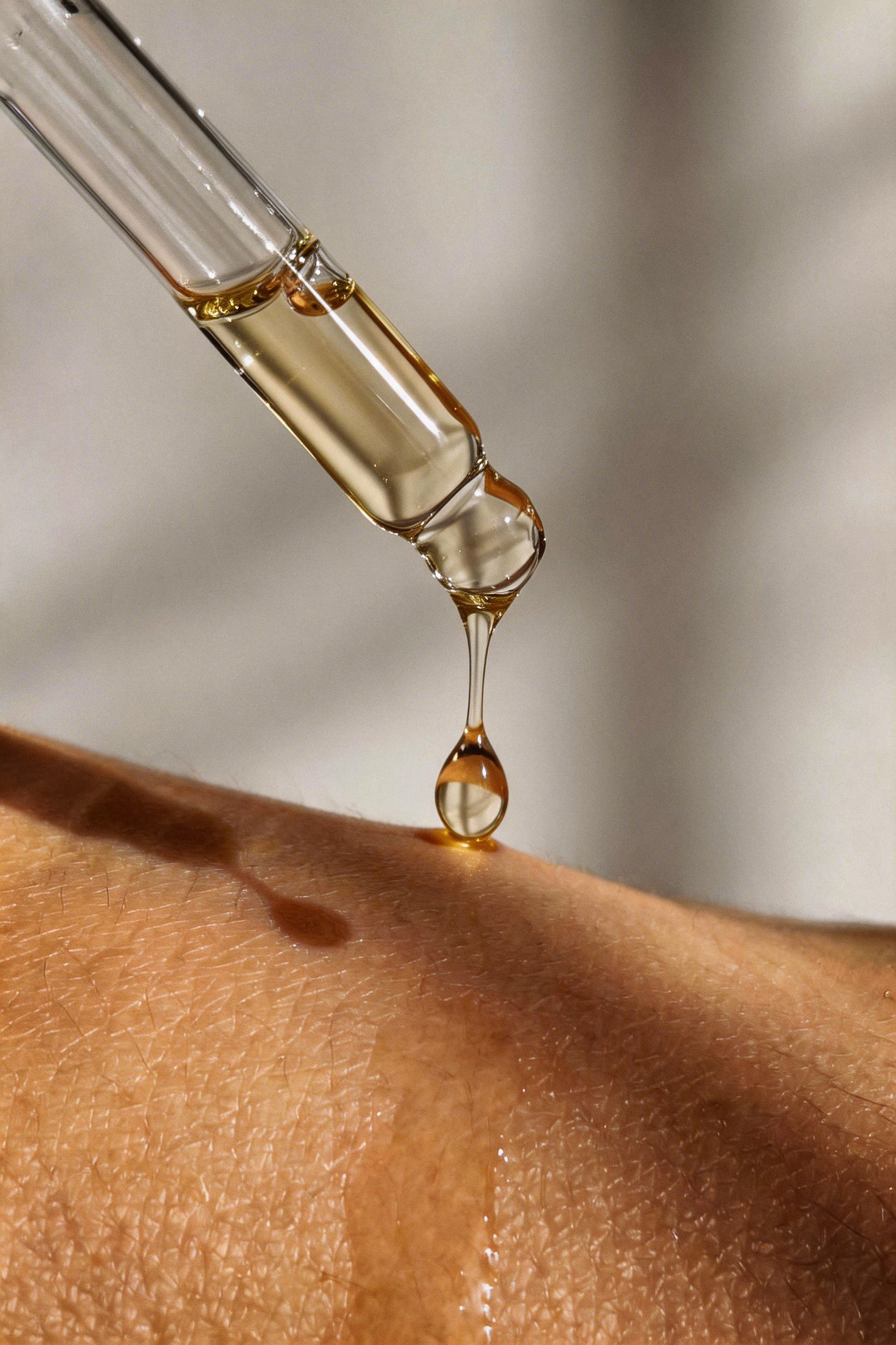 A close-up of a clear glass dropper releasing a single golden liquid drop onto human skin, with soft shadows and a warm neutral background.
