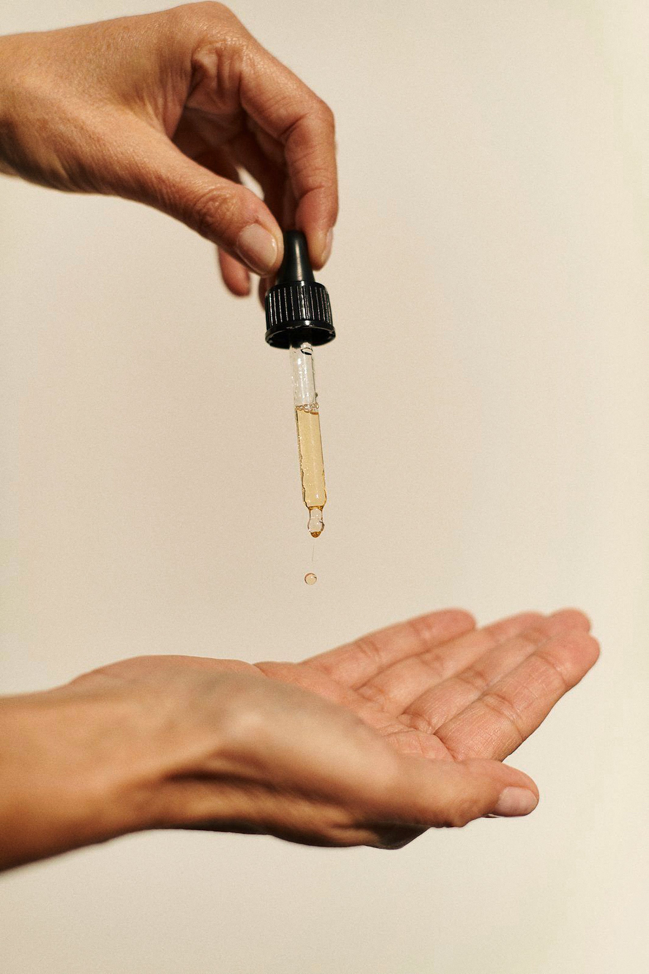 A close-up of a hand holding a glass dropper above another open hand, with a single drop of golden liquid hovering between them against a plain light beige background.