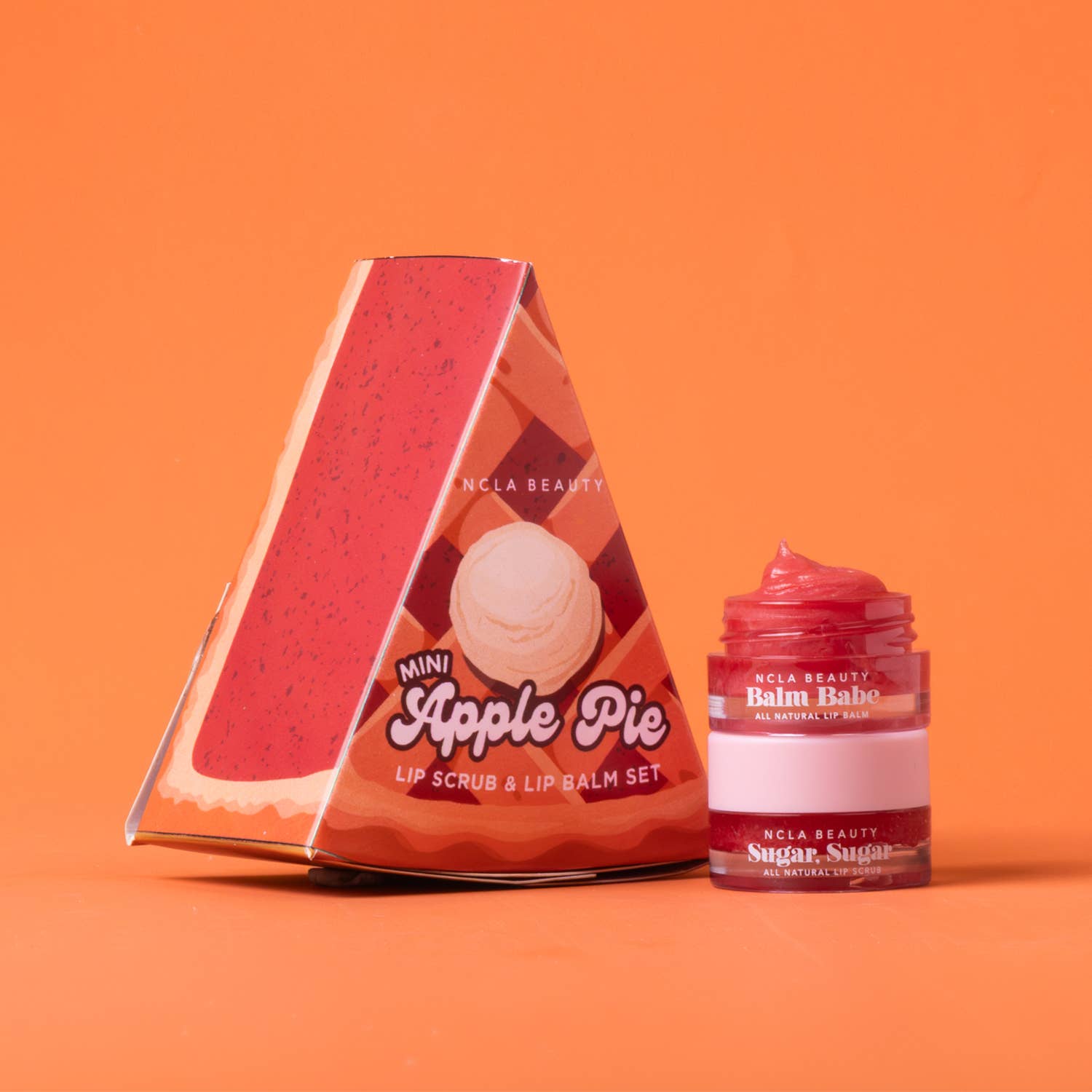 NCLA Beauty Mini Apple Pie lip scrub and lip balm set, featuring a triangular cardboard box with an apple pie illustration and a small jar of pink balm beside it, set against a solid orange background.