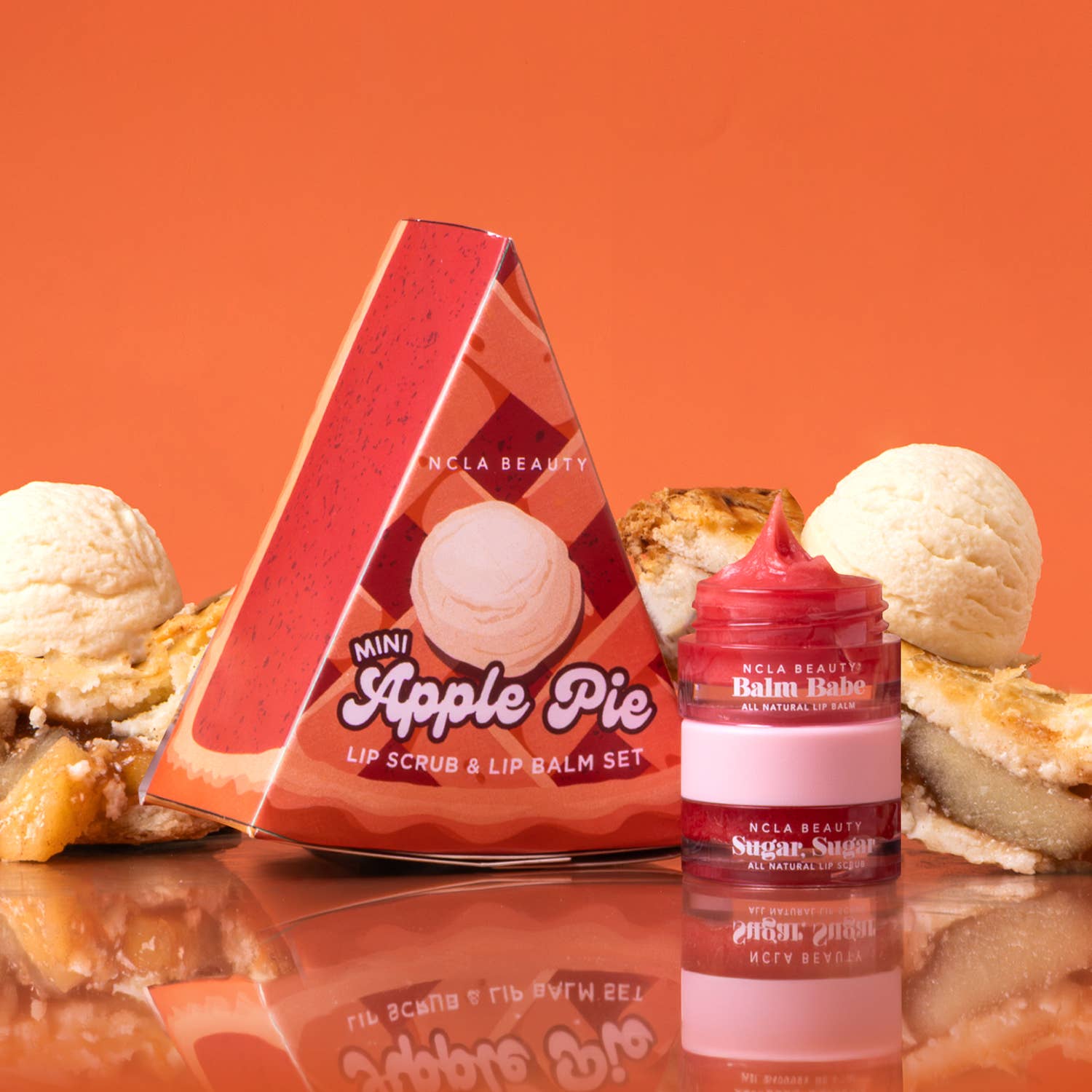 NCLA Beauty Mini Apple Pie lip scrub and lip balm set, featuring a red triangular box and a matching red jar labeled 'Sugar Sucker' on a reflective surface with apple pie and ice cream props against an orange background.