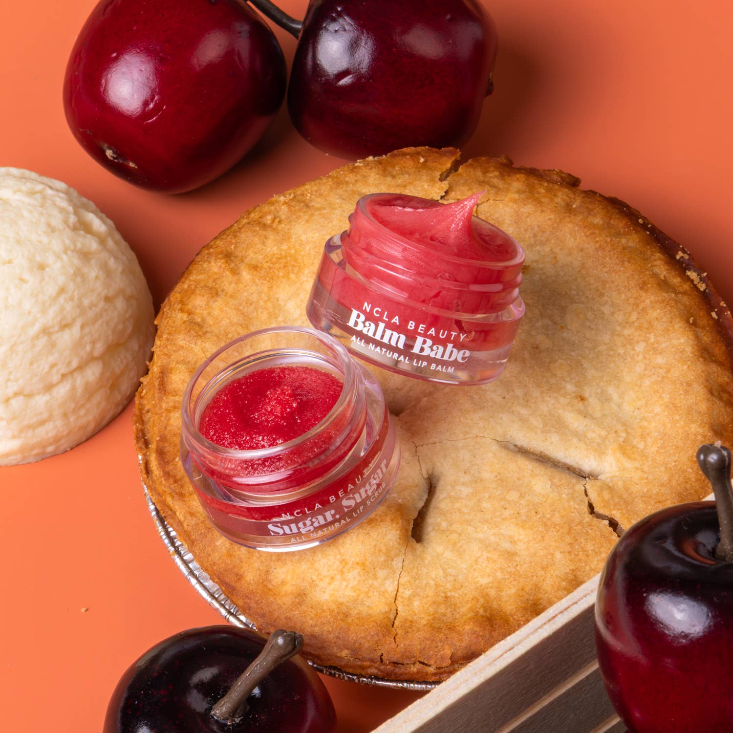 Two small jars of BCGA Beauty 'Balm Babe' sugar mask balm, one with a pinkish-red balm and one with a bright red glittery balm, styled on a golden-brown pie crust with whole cherries and a scoop of vanilla ice cream on an orange background.