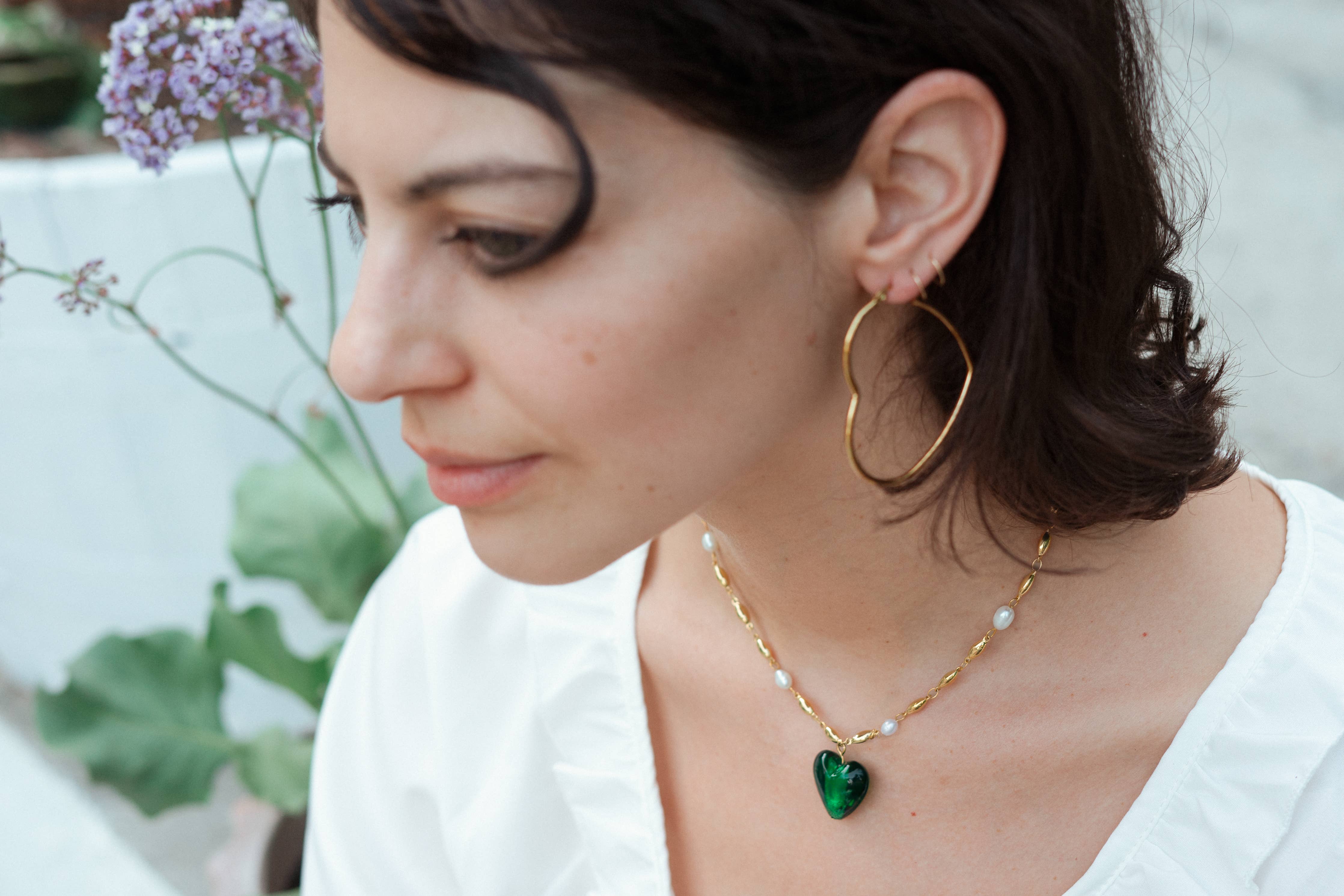 A close-up lifestyle shot of a woman wearing a gold chain necklace with a green heart-shaped pendant and matching gold hoop earrings.