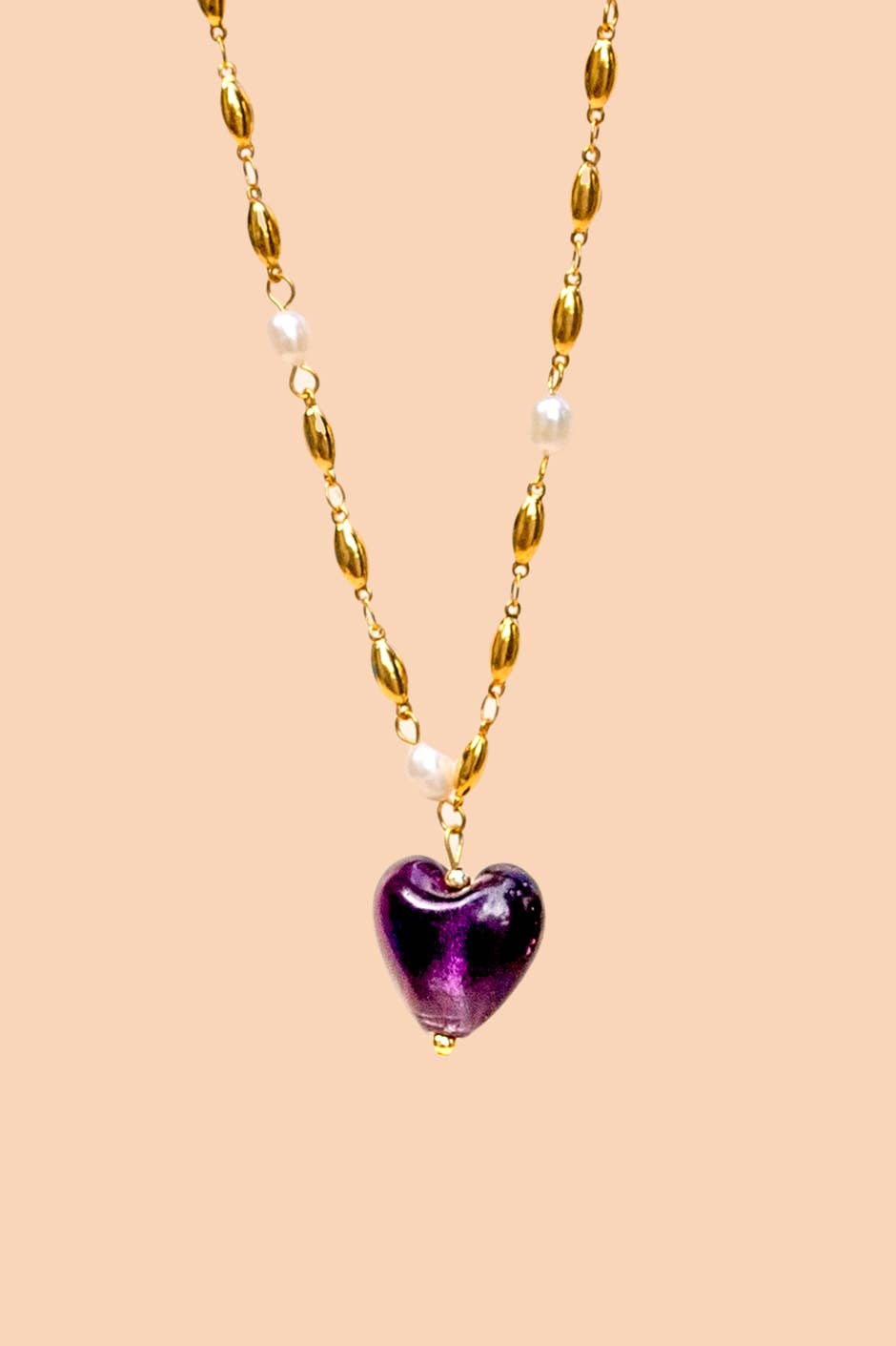 A gold chain necklace featuring a glossy purple heart-shaped pendant and small white pearl-like beads, displayed against a solid pale peach background.