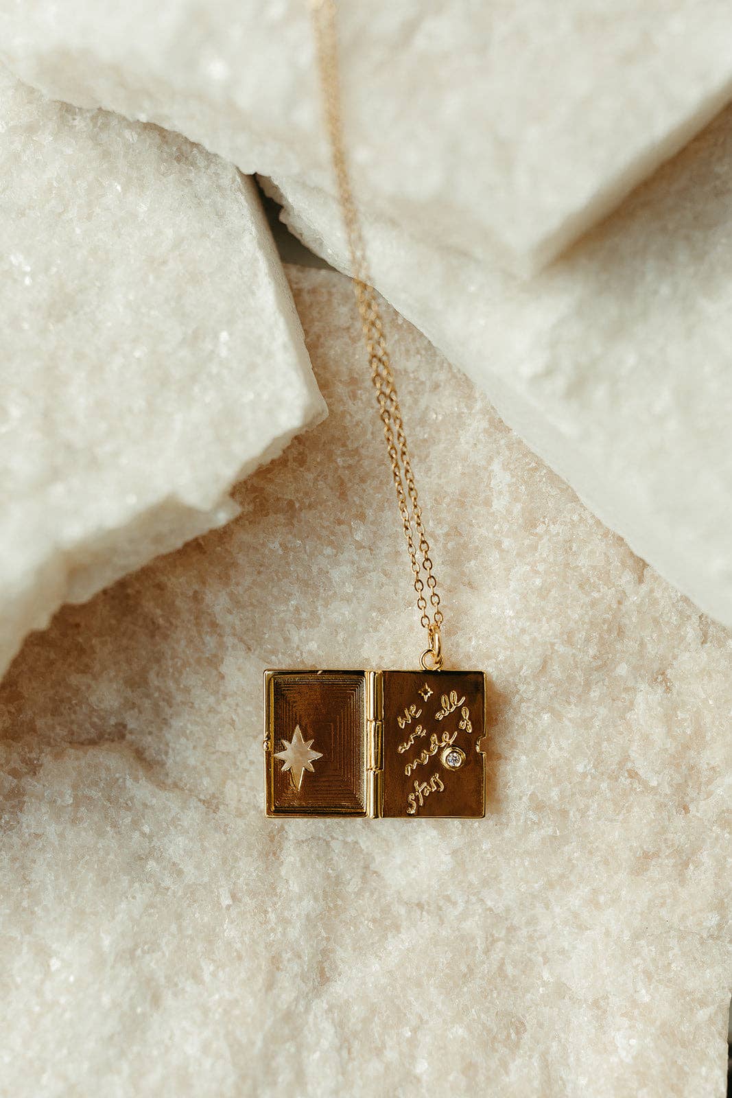 A gold-toned locket pendant necklace resting on textured white stone. The open locket features one side with a star design and the other with cursive text and a small clear gemstone.