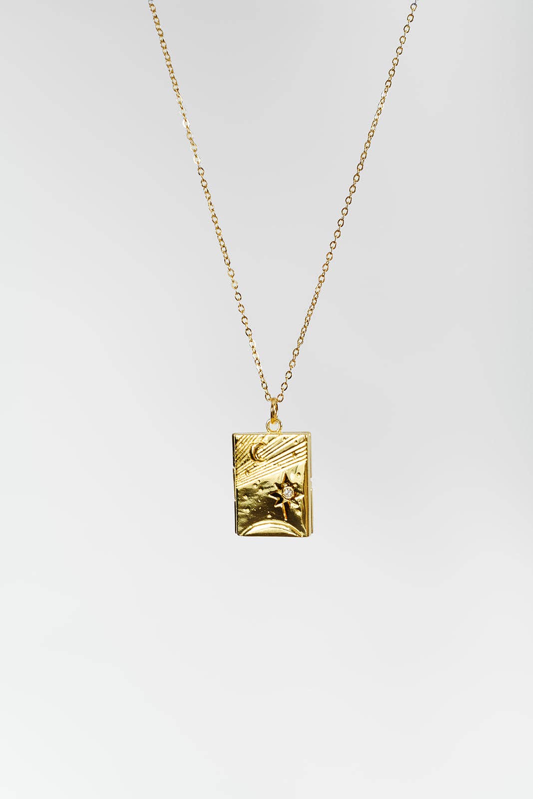 A gold pendant necklace featuring a rectangular, textured charm with a star motif, suspended from a delicate gold chain against a light gray background.