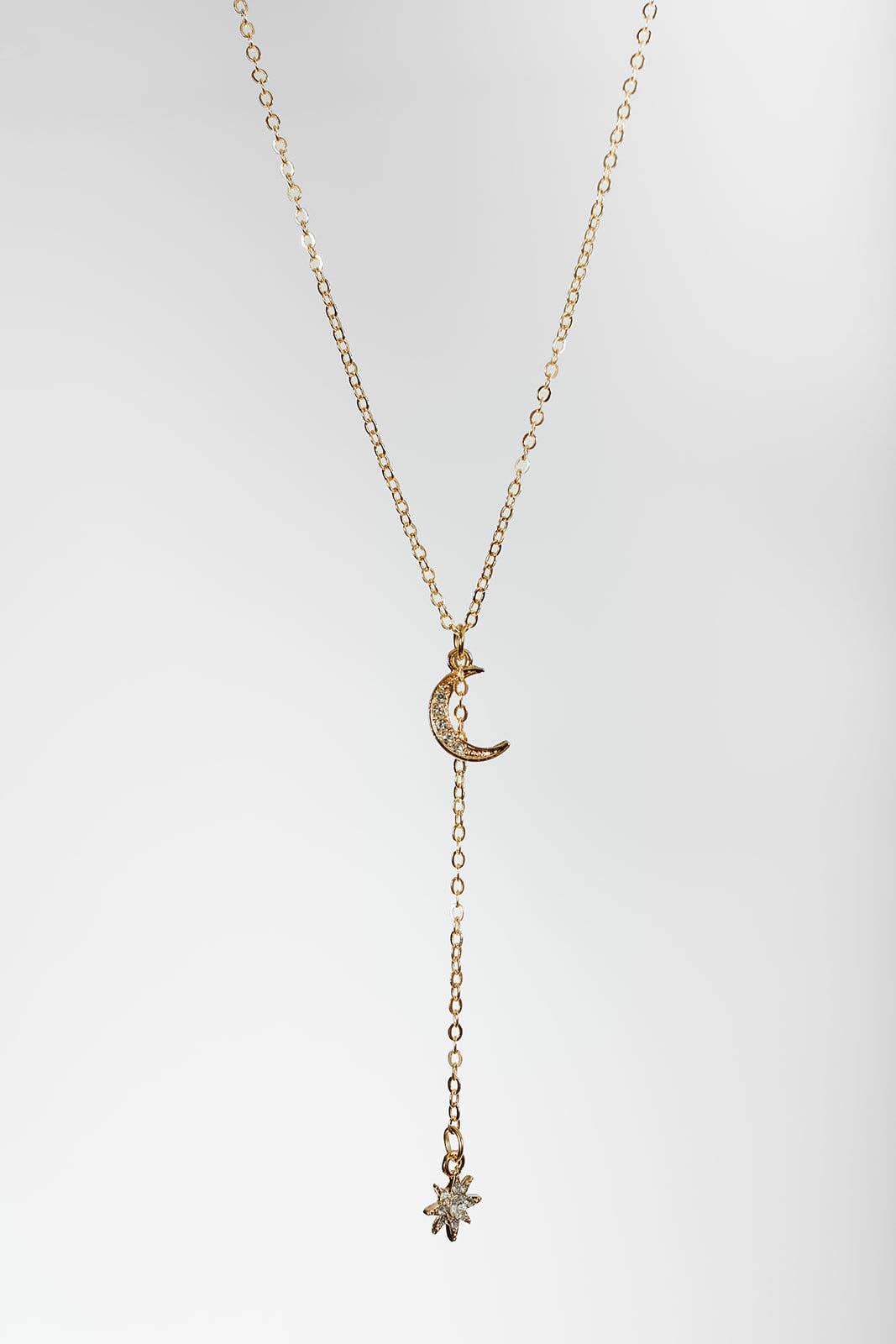 A delicate gold necklace featuring a crescent moon pendant adorned with small clear stones, with a long drop chain ending in a starburst charm, set against a light gray gradient background.