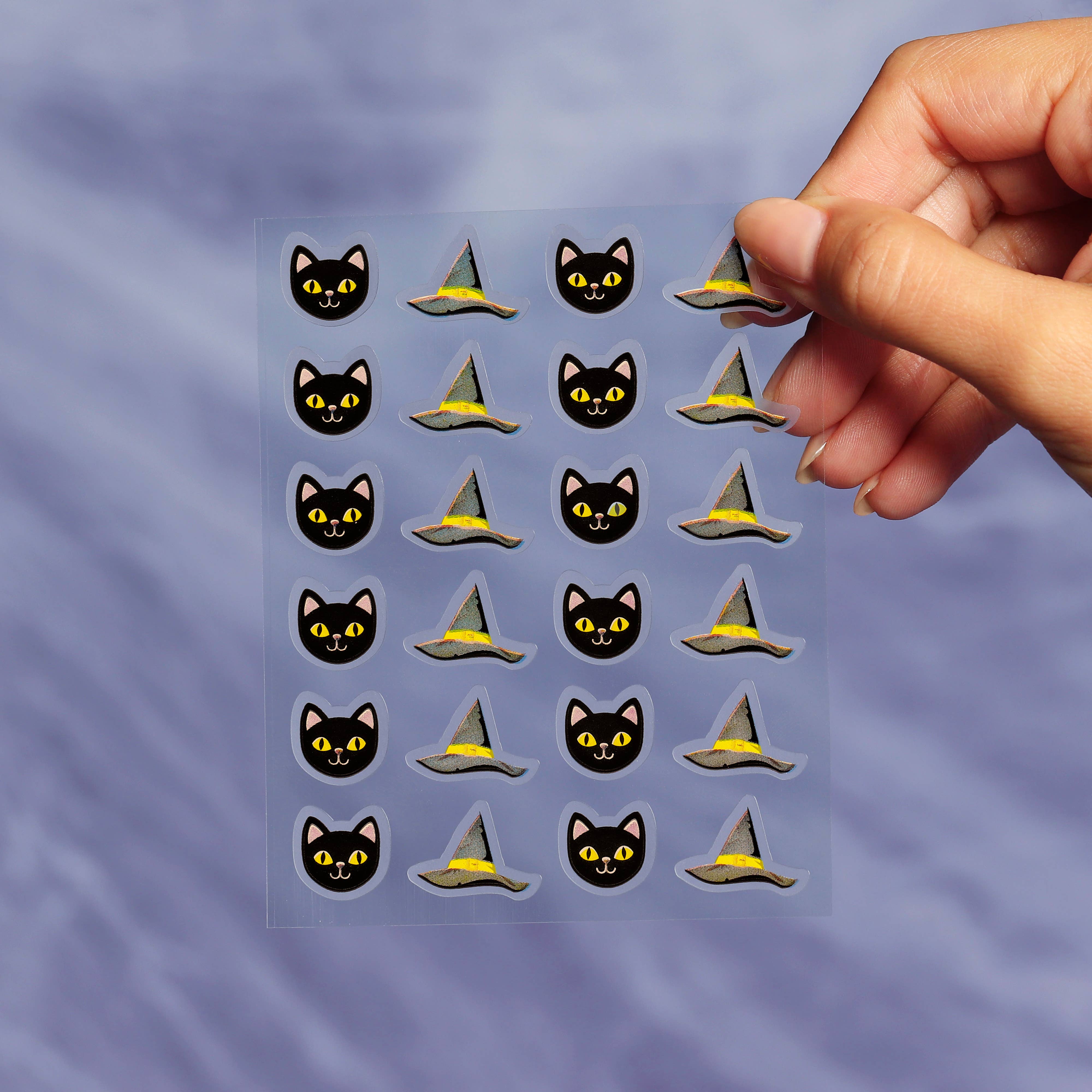 Sheet of Halloween-themed stickers with cats and witch hats held by a hand against a purple background.