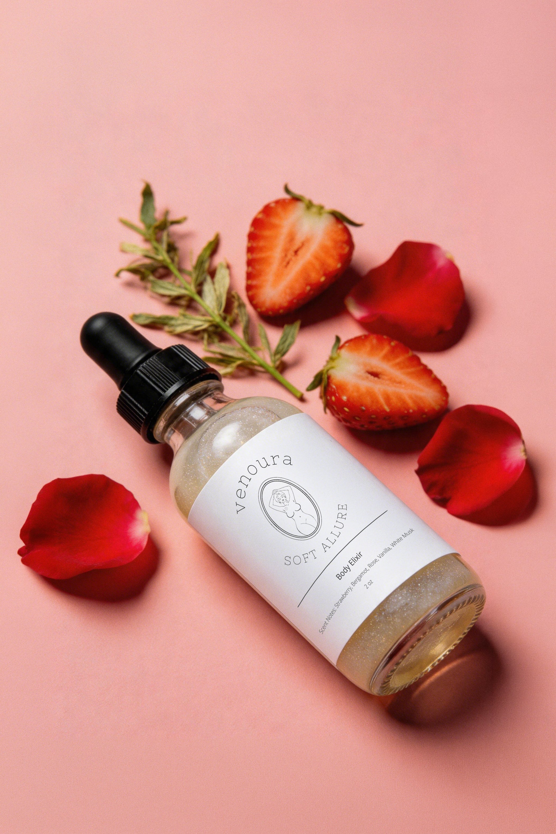 A glass dropper bottle of Venoura Soft Alchemy body butter with a white label and black dropper cap, styled on a pink background with strawberry slices, rose petals, and a sprig of rosemary.