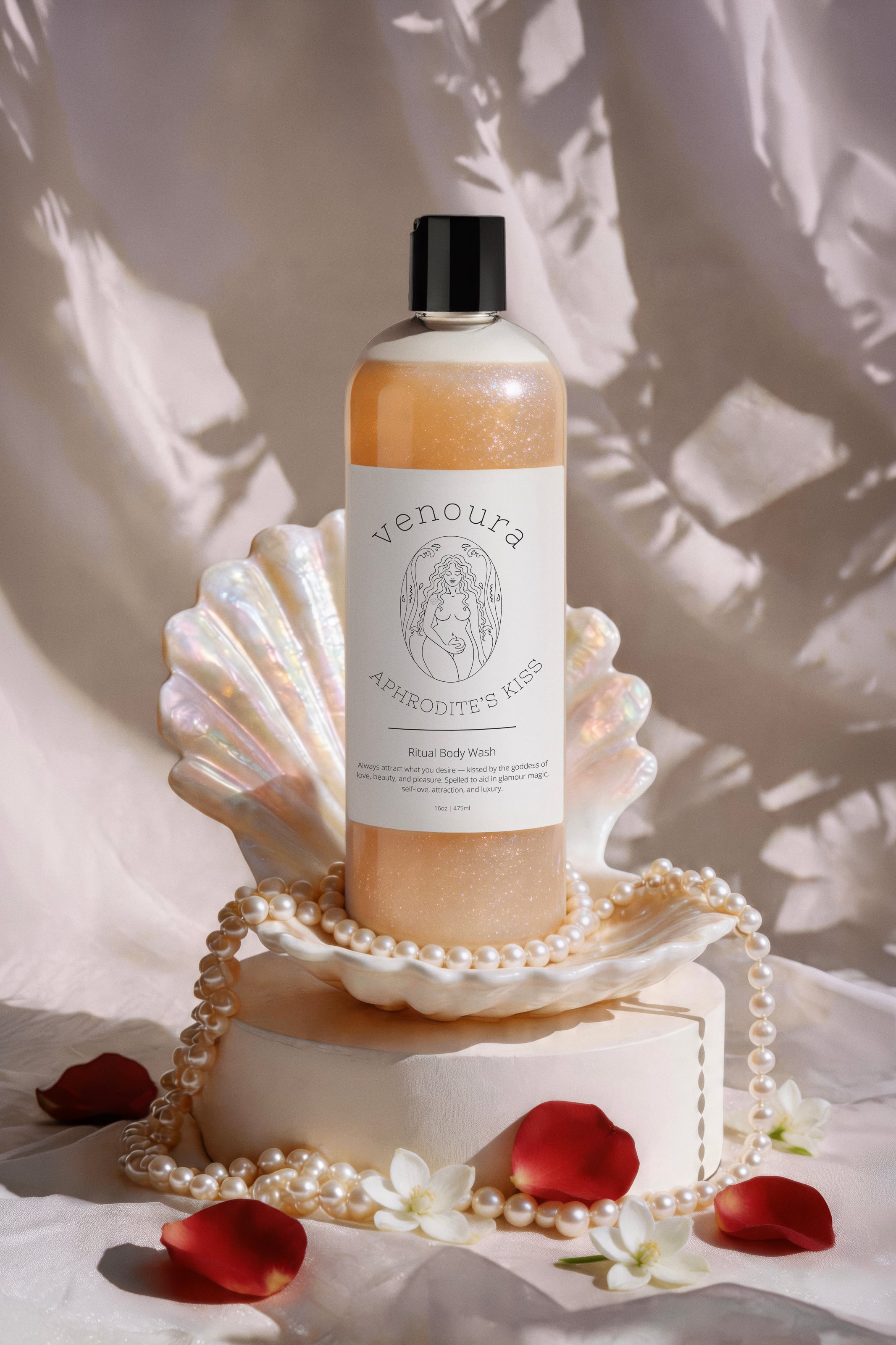 Venoura Aphrodite's Kiss Body Wash in a clear bottle with a black cap, styled on a white shell pedestal with pearls, red rose petals, and white flowers against a draped beige fabric background.