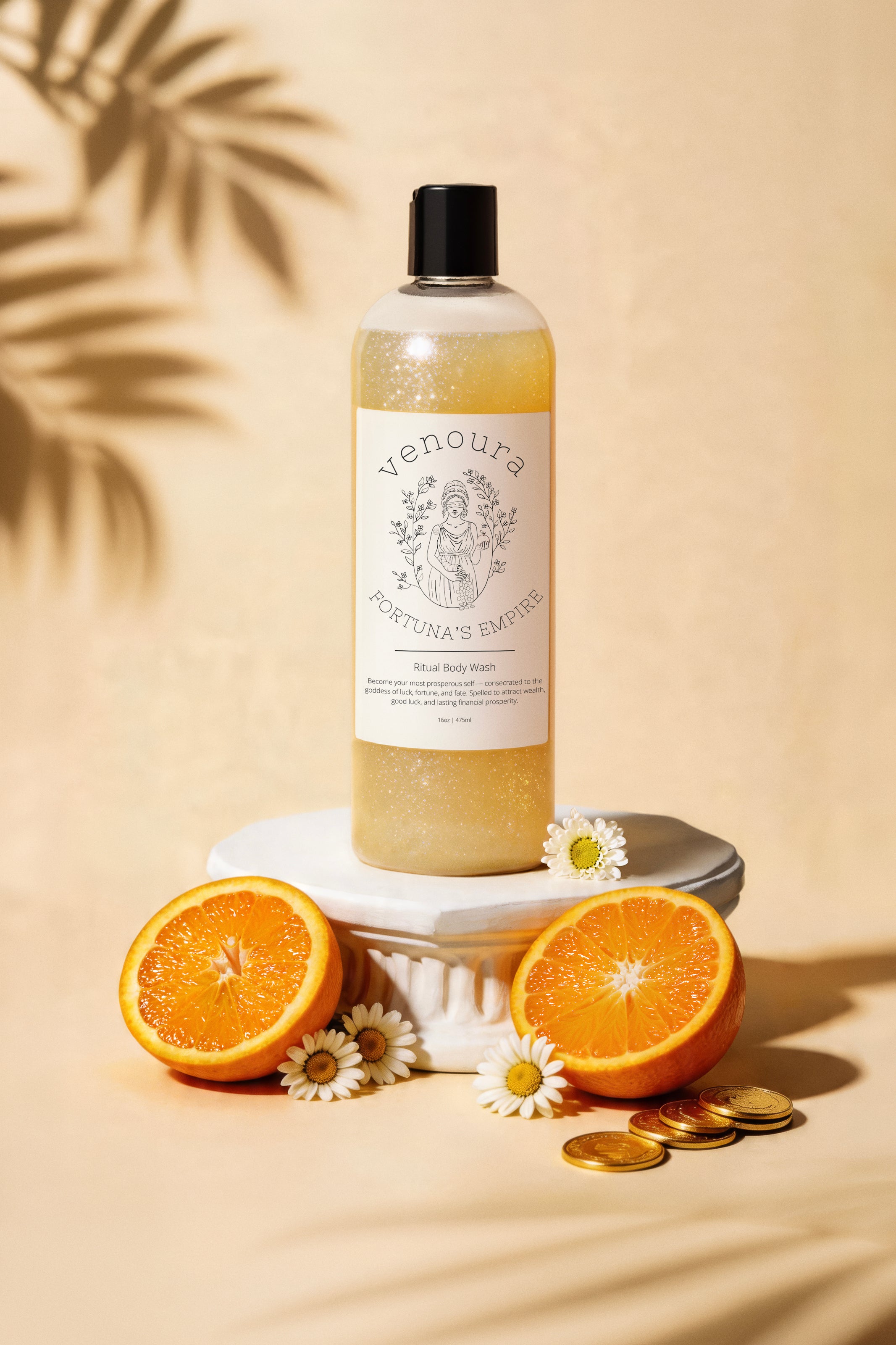 Venoura Fortuna's Empire Body Wash in a clear bottle, styled on a white pedestal with orange slices, chamomile flowers, and gold coins on a tan background with leaf shadows.