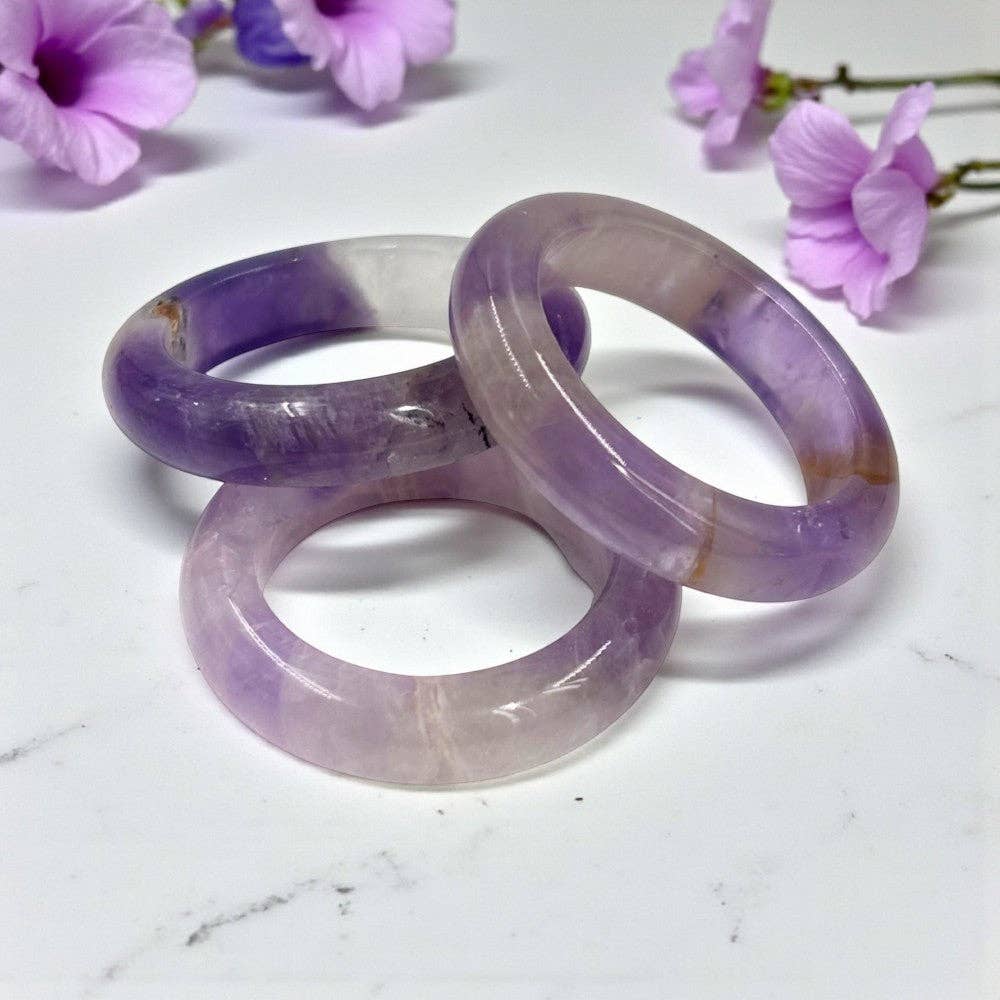 Three purple and clear bangles on a white surface with flowers in the background