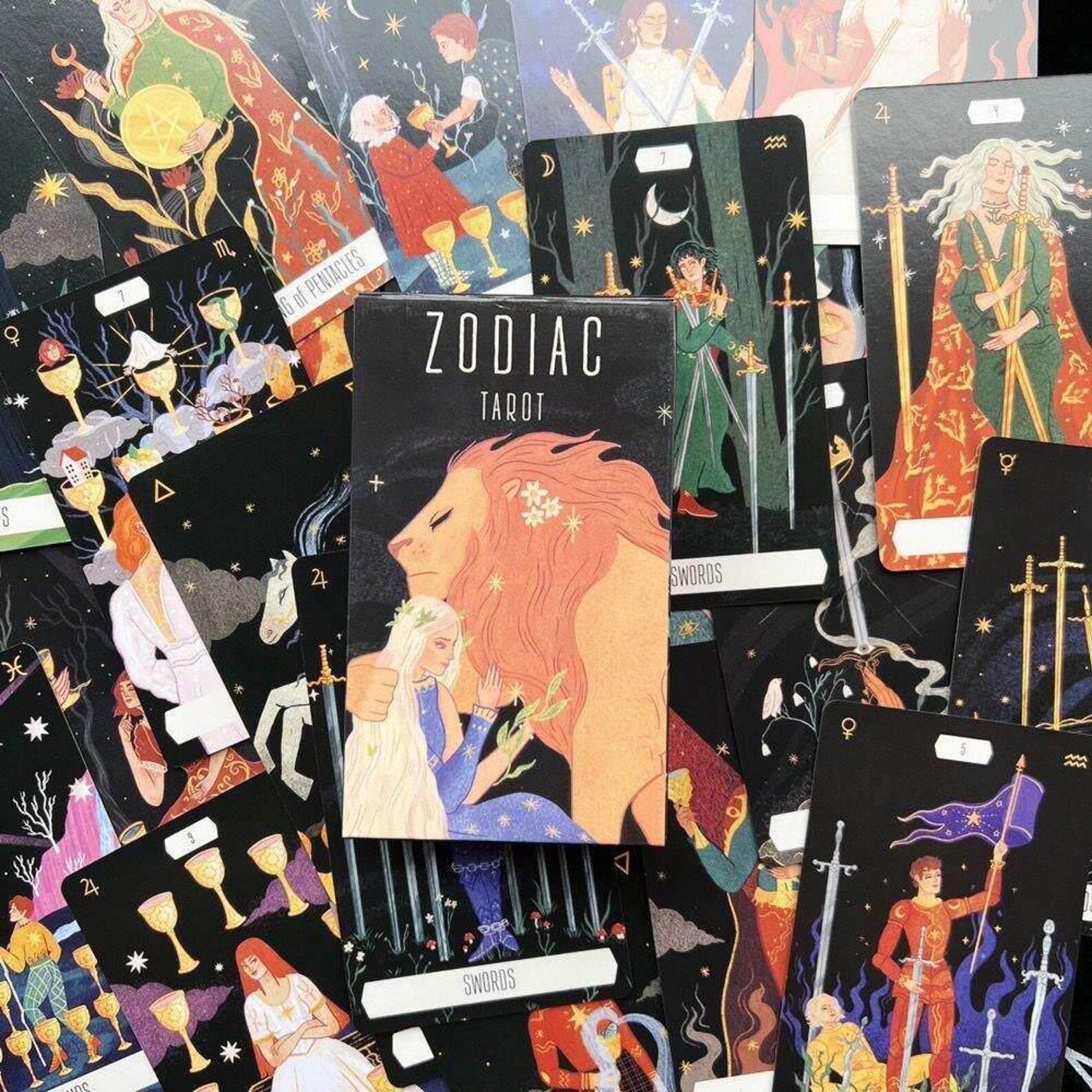 A boxed tarot deck titled 'ZODIAC TAROT' is centered among several illustrated tarot cards spread out on a dark surface. The box art shows a lion and a woman, and the surrounding cards display other zodiac-themed illustrations.
