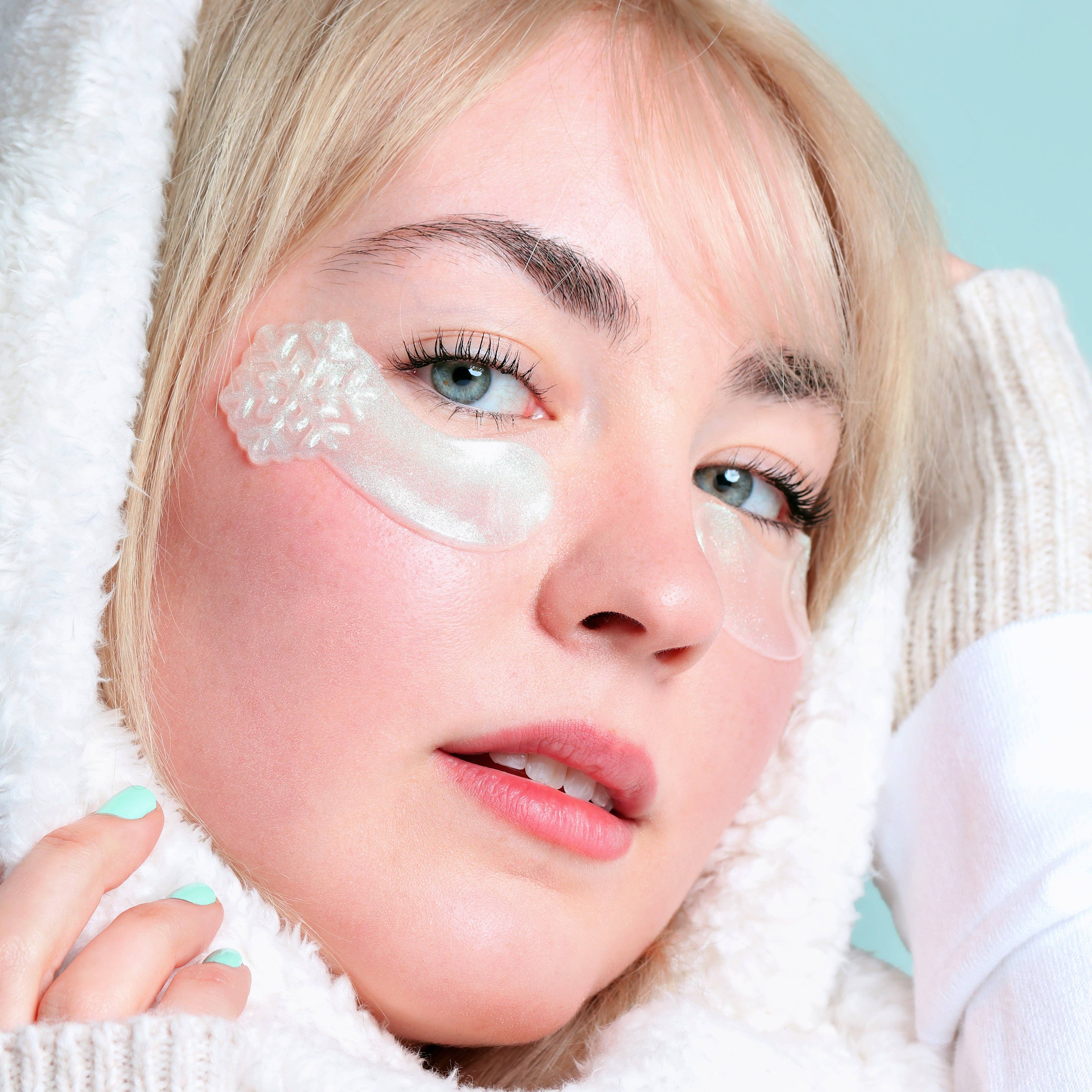 A close-up of a blonde woman wearing clear under-eye patch masks on her eyes, wrapped in a white fleece blanket against a light blue background.