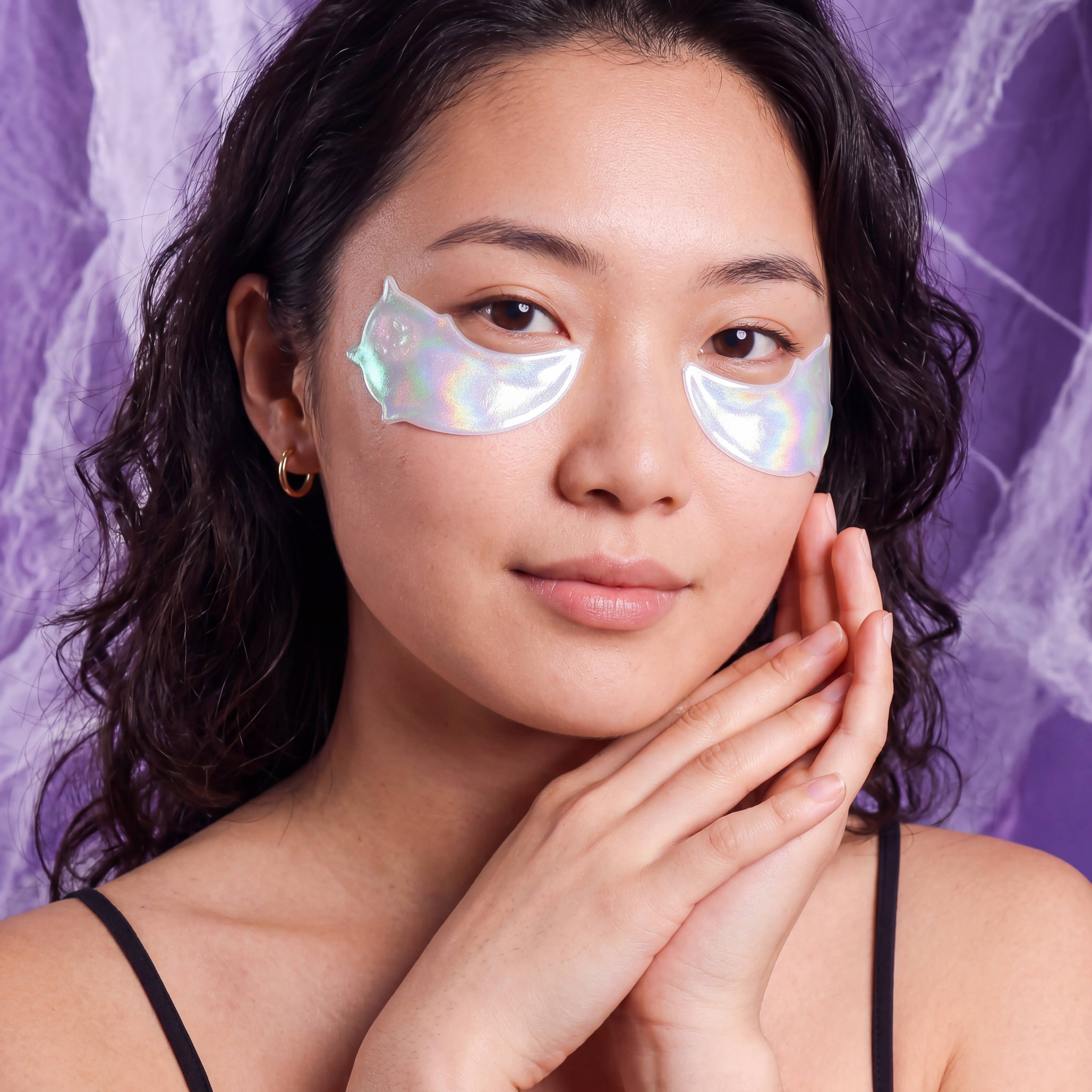 A woman with dark curly hair wearing iridescent cat-shaped eye masks under her eyes, posing against a purple marbled background.