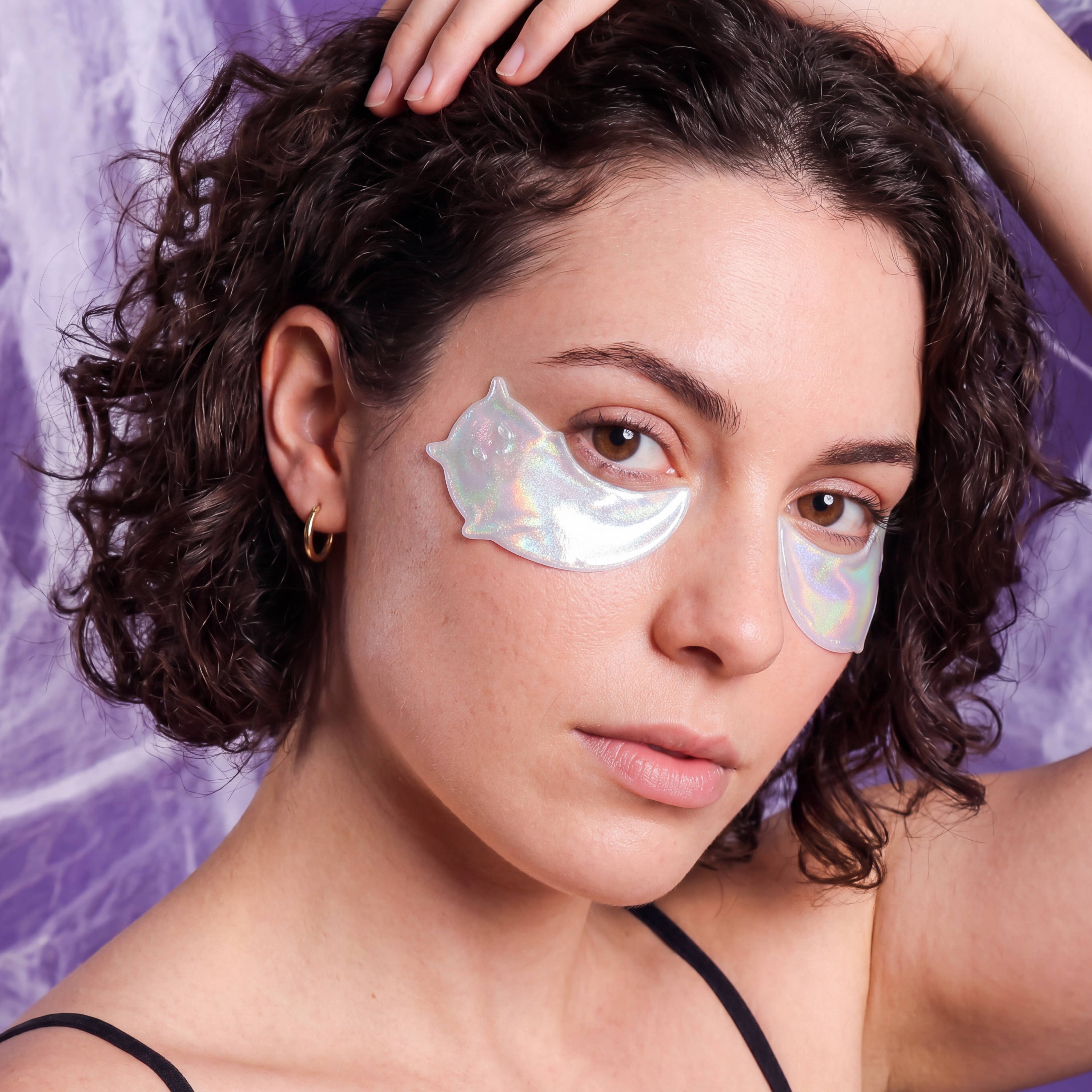 A close-up of a woman with curly dark hair wearing two iridescent, semi-translucent under-eye patches against a purple marbled background.