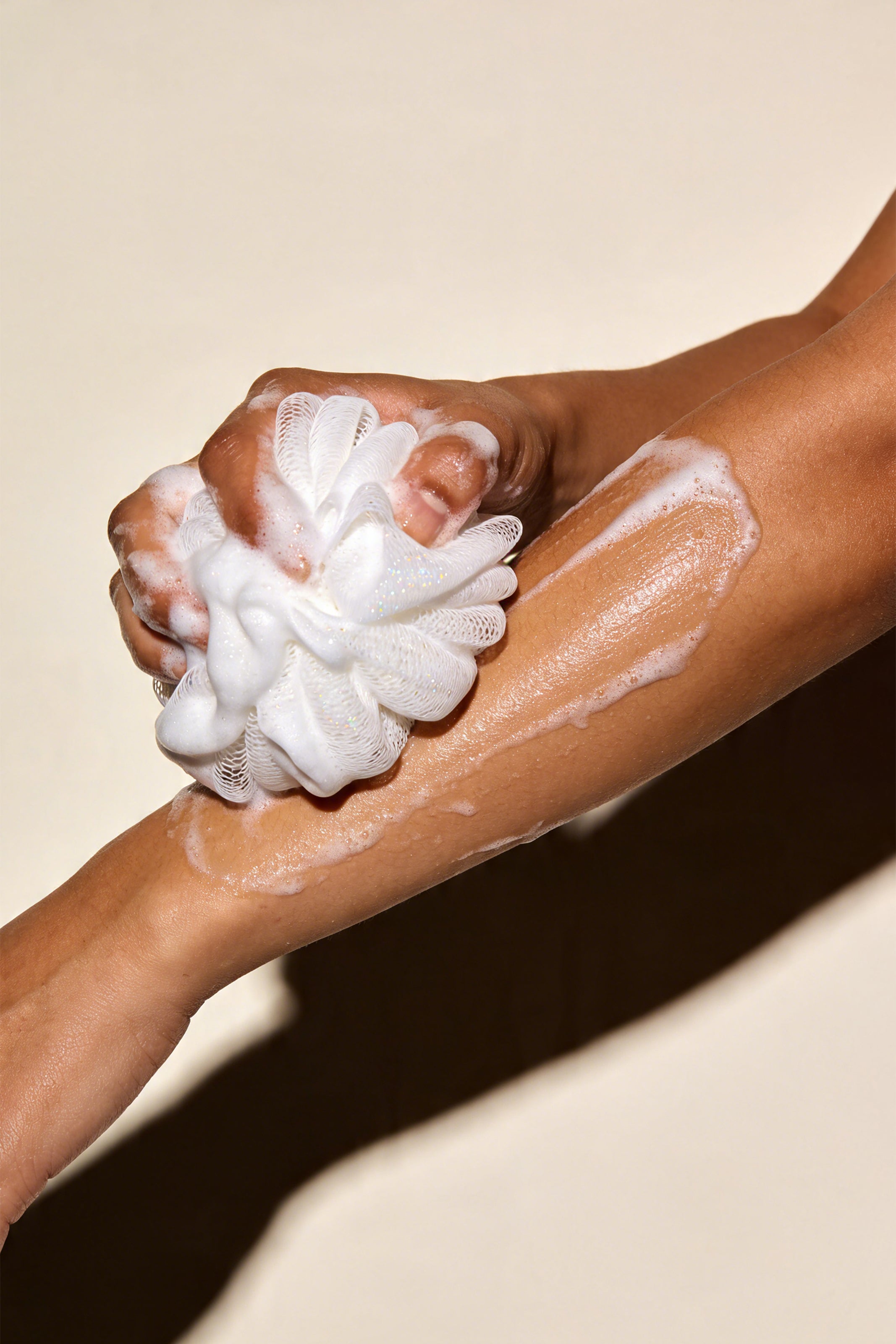A close-up of a person's forearm and hand being lathered with Venoura's Aphrodite's Kiss Body Wash white soap foam while holding a white textured scrubber or loofah against a warm beige background.