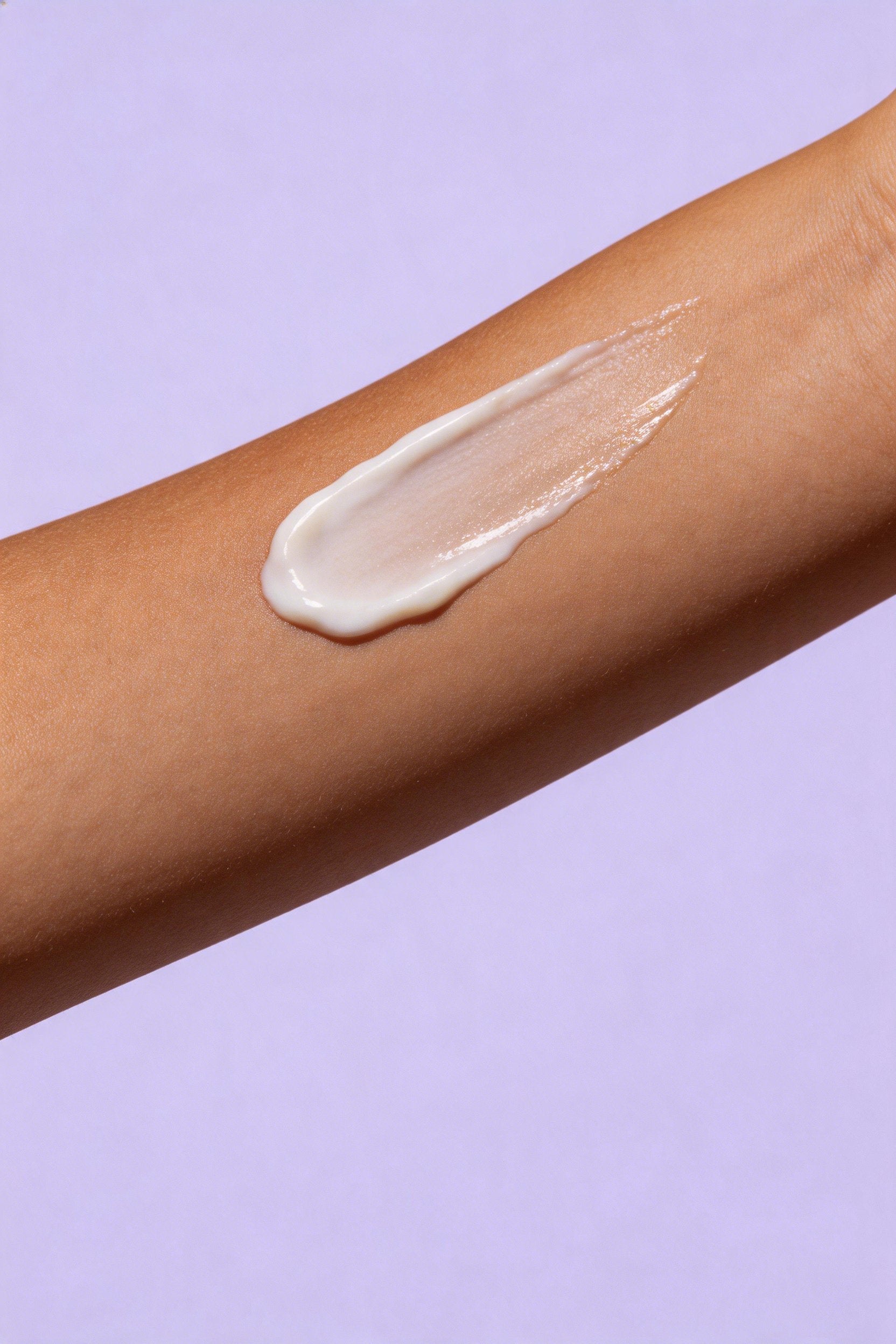 A thick, creamy off-white lotion or serum swatch applied in a diagonal smear across a person's forearm, set against a solid light purple background.