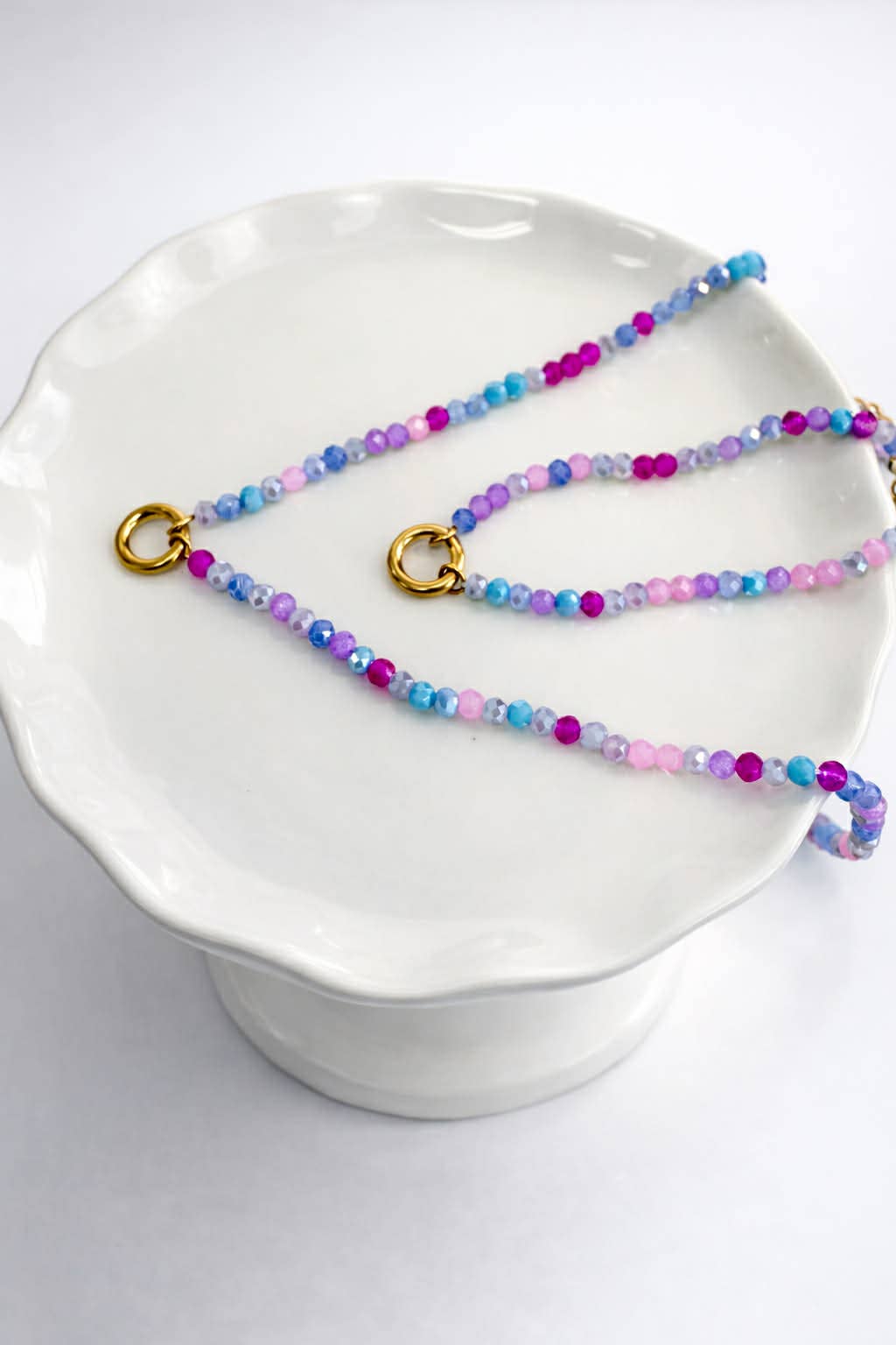 A beaded necklace with gold-toned rings, featuring pastel blue, pink, and purple faceted beads, displayed on a white ceramic pedestal.