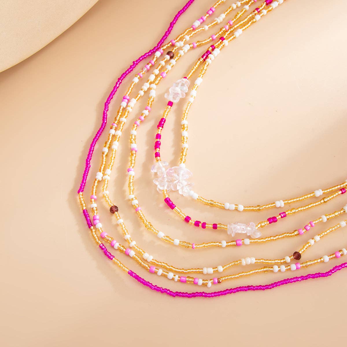 A close-up of a multi-strand beaded necklace draped on a warm beige surface. The necklace features metallic gold seed beads, bright magenta pink beads, small white pearl-like beads, and a few clear faceted crystal beads.