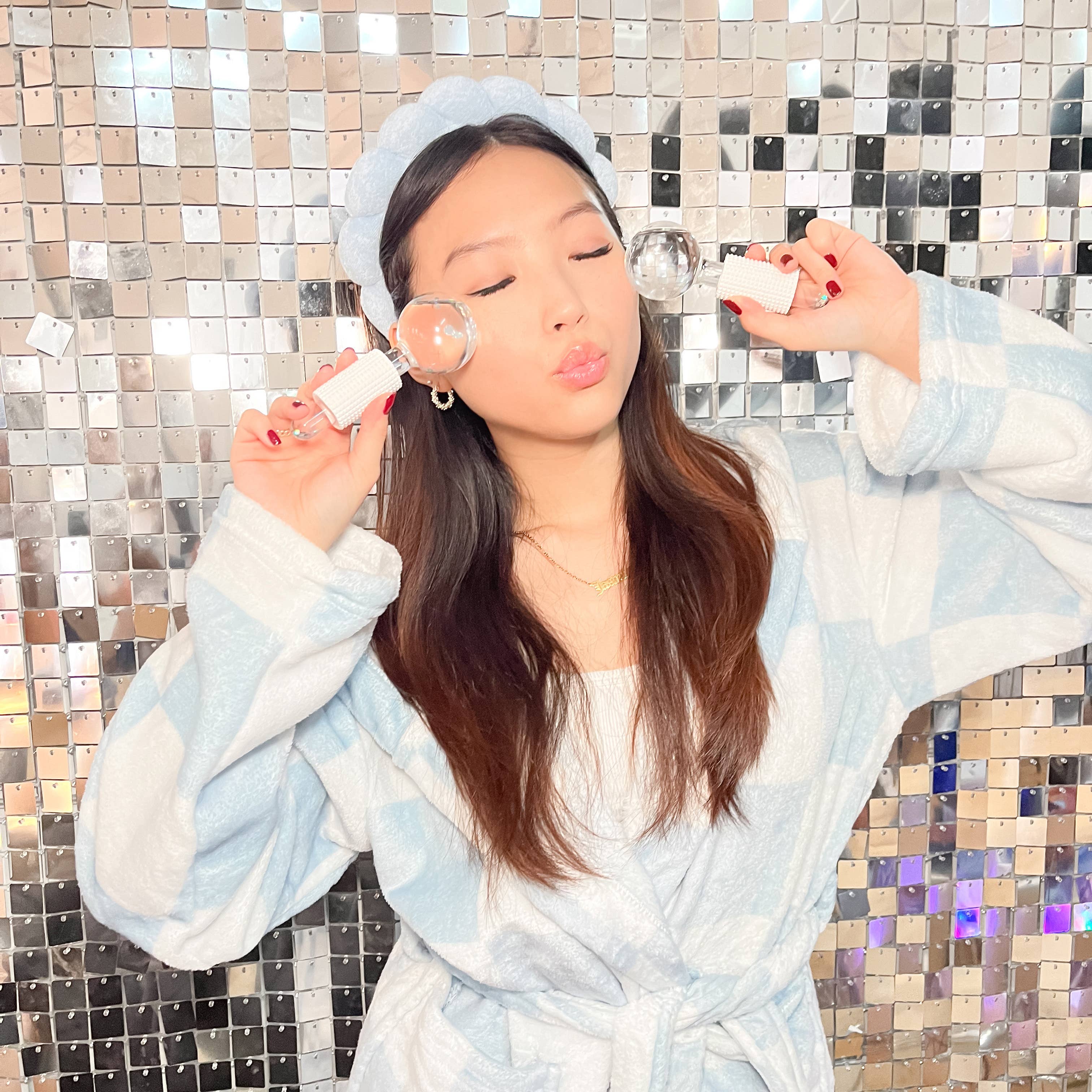 Person in a light blue robe holding skincare products against a mosaic tile wall.